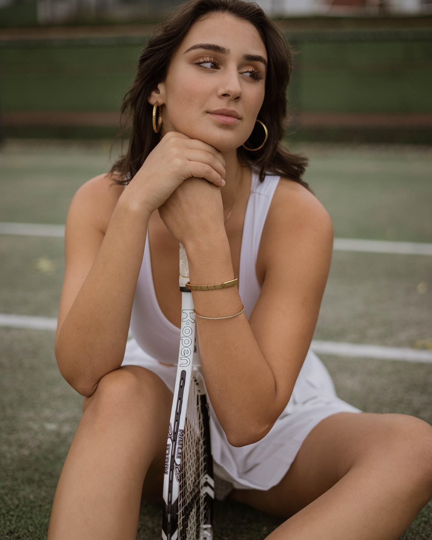 Brunette woman in white tennis dress sits on court holding racket with gold hoop earrings.