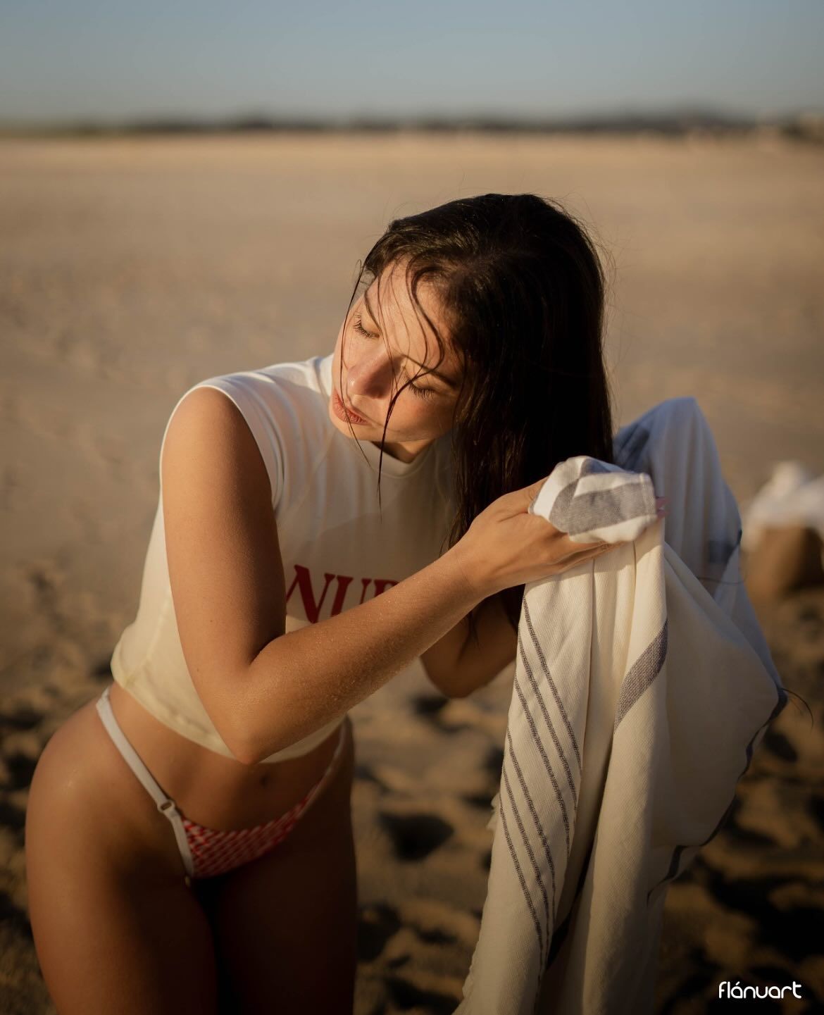 Dark-haired woman in white top and bikini drying off with towel on sandy beach.