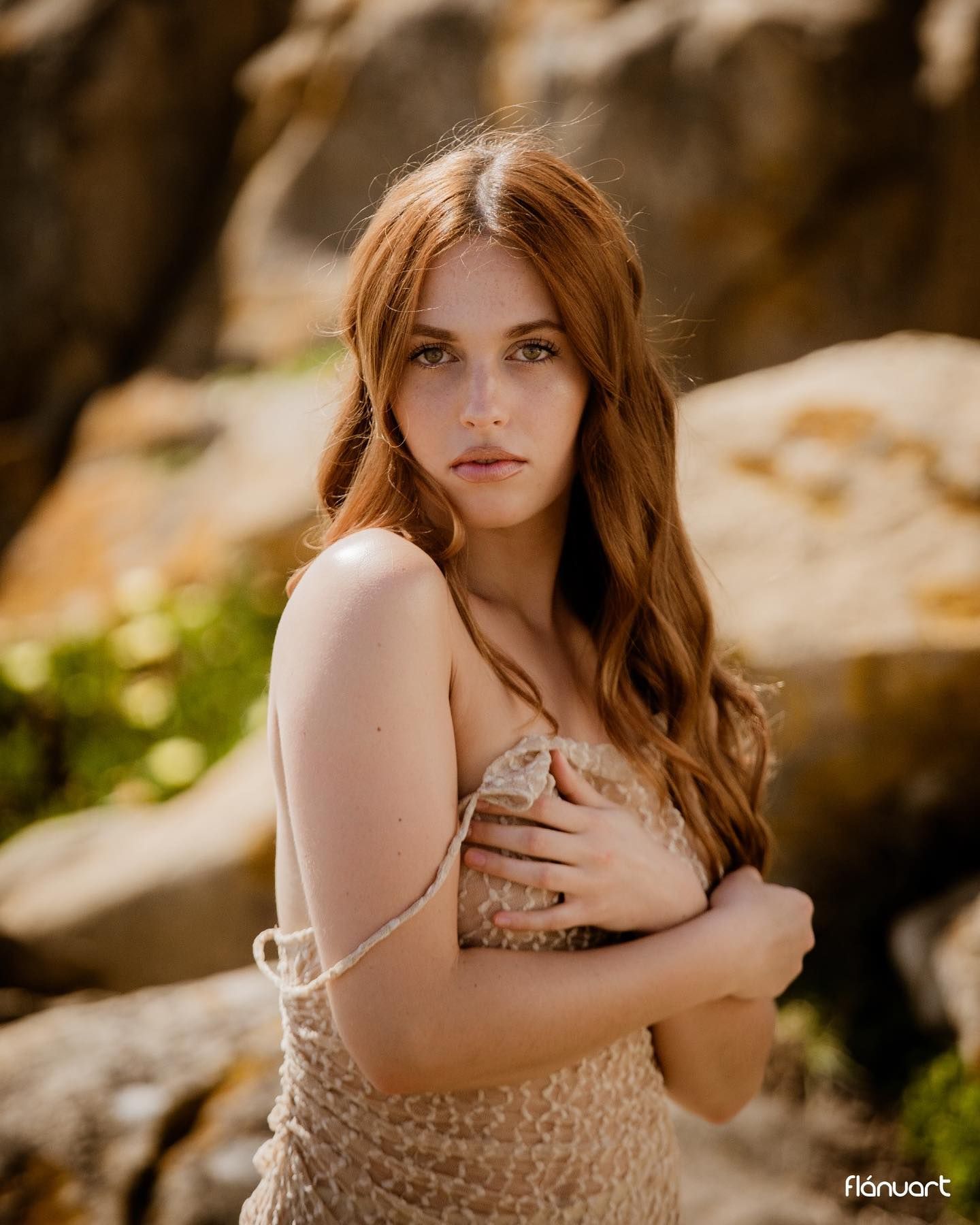 Young woman with long red hair in lace dress posing against rocky outdoor backdrop.