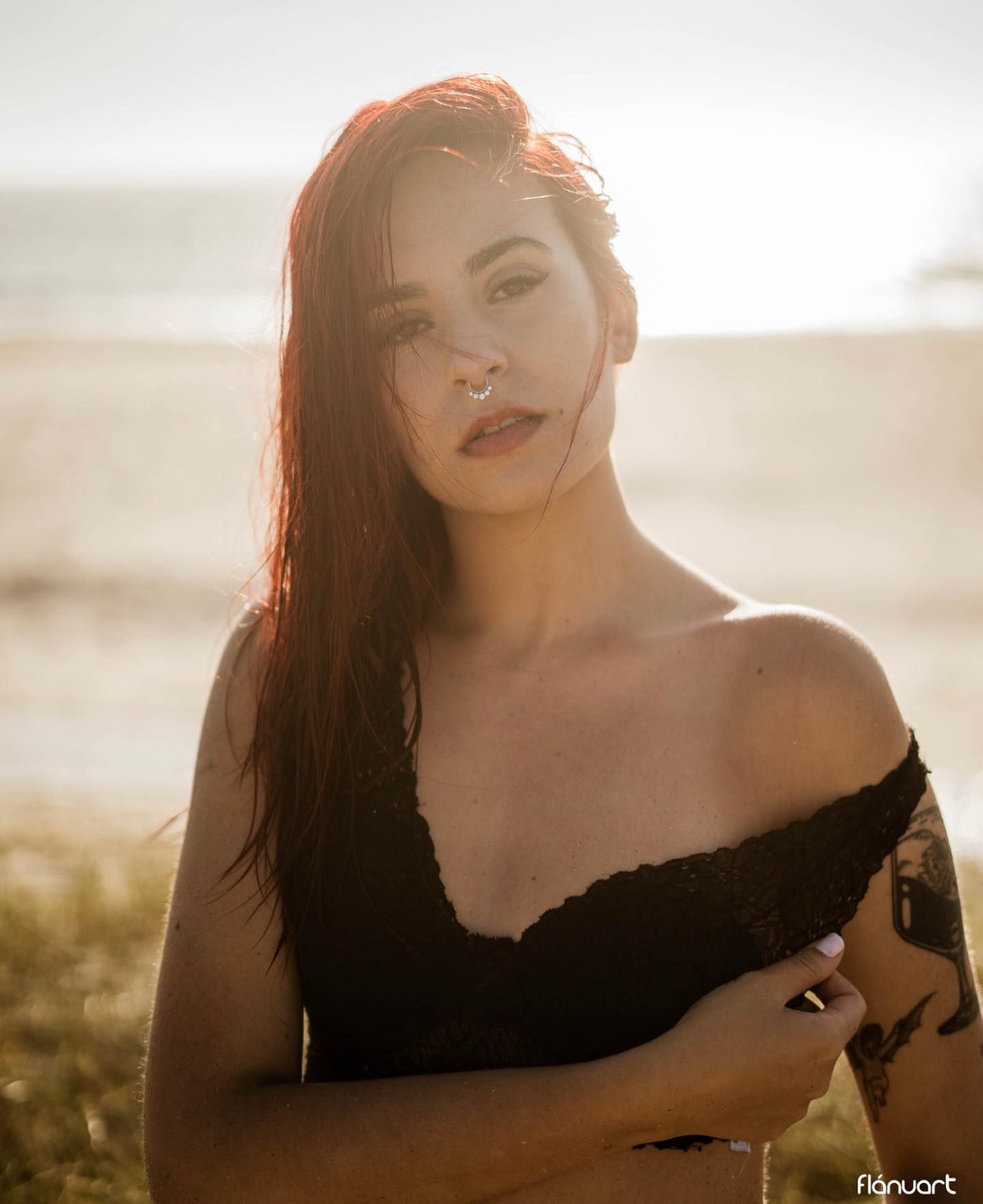 Redhead woman in black off-shoulder top poses on beach with golden backlight.