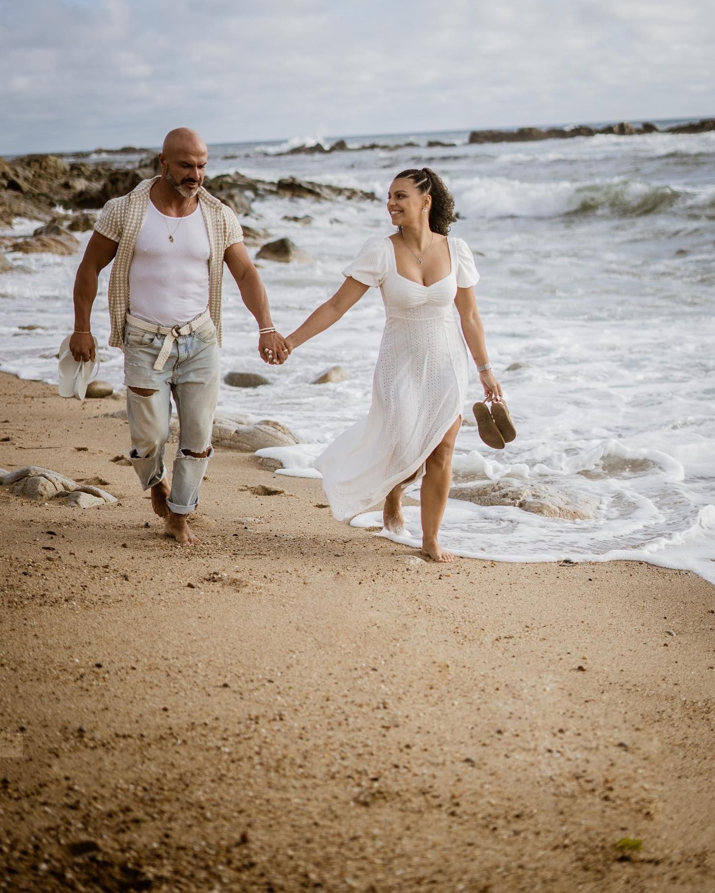 Couple walking hand-in-hand barefoot along sandy beach with ocean waves, woman in white flowing dress.