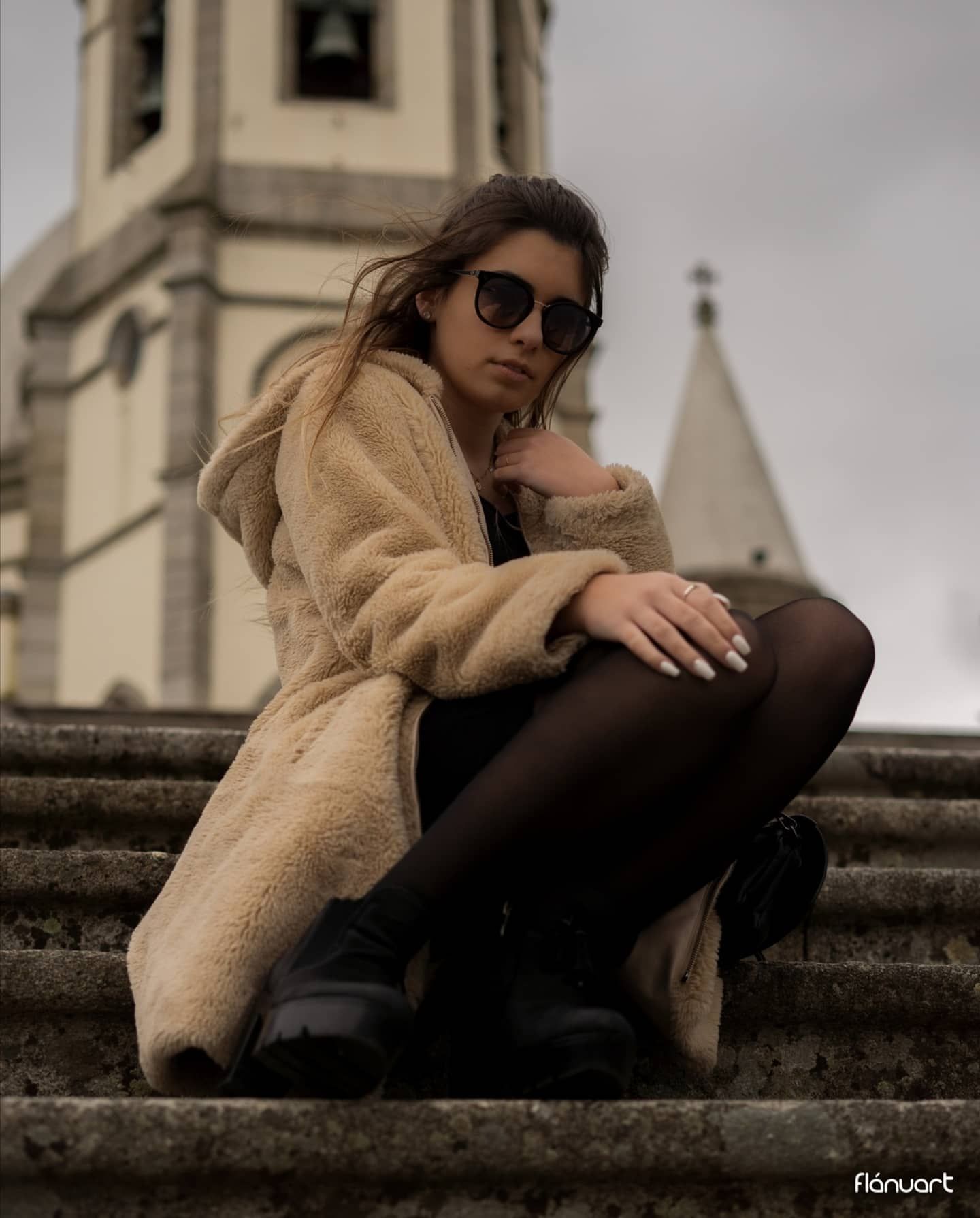 Woman in beige teddy coat and sunglasses sits on stone steps before a historic church.