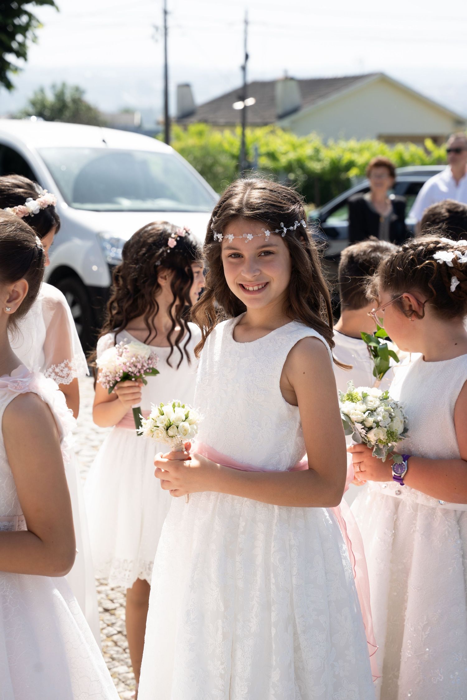 Young girls in white dresses holding bouquets at a First Communion ceremony, smiling outdoors.