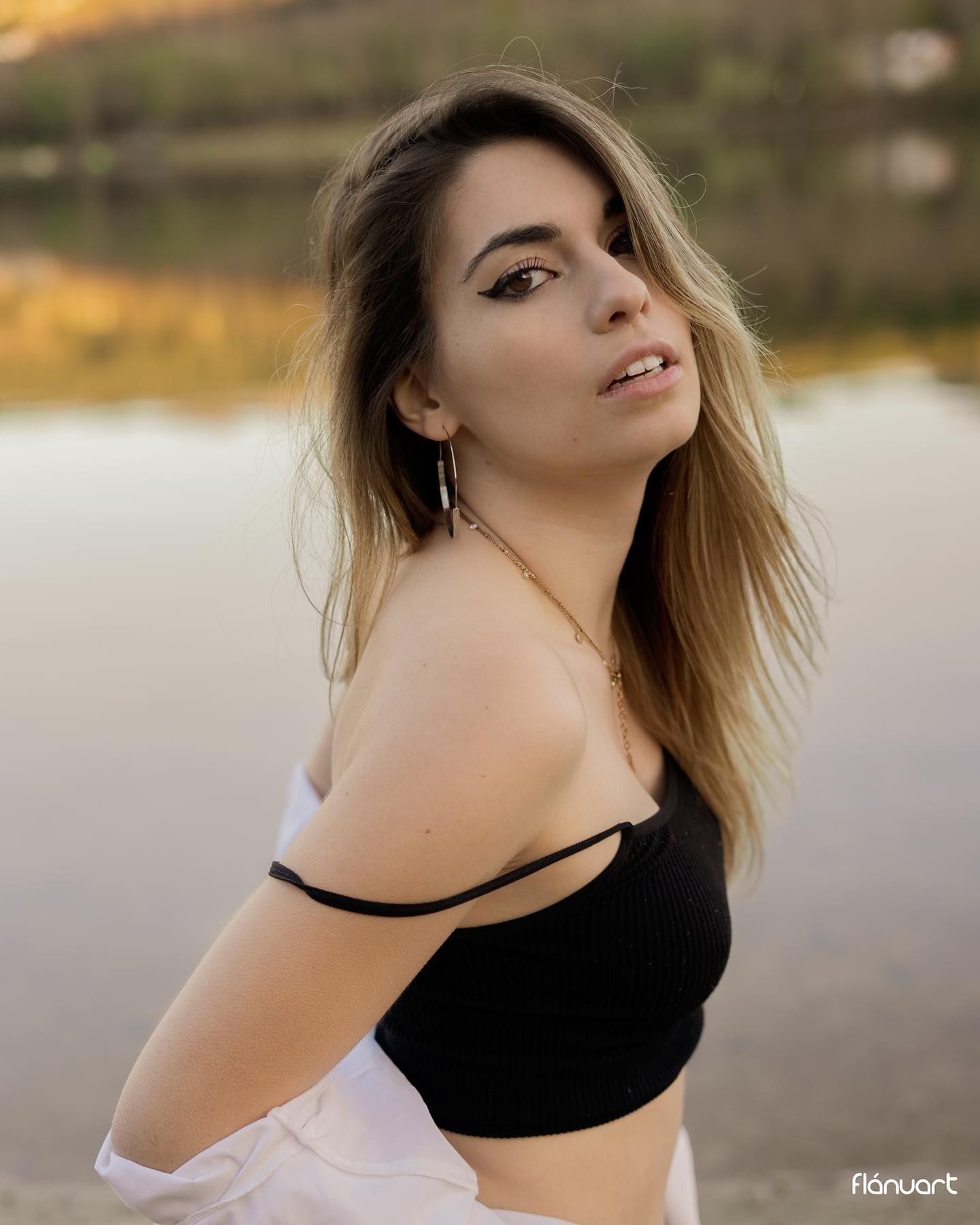Young woman in black crop top poses by a lakeside at golden hour with hair blowing softly.