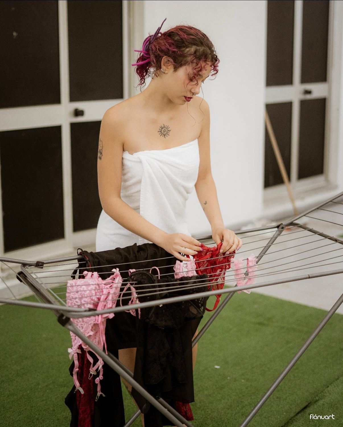 Woman with pink hair wrapped in white towel hanging laundry on outdoor drying rack.