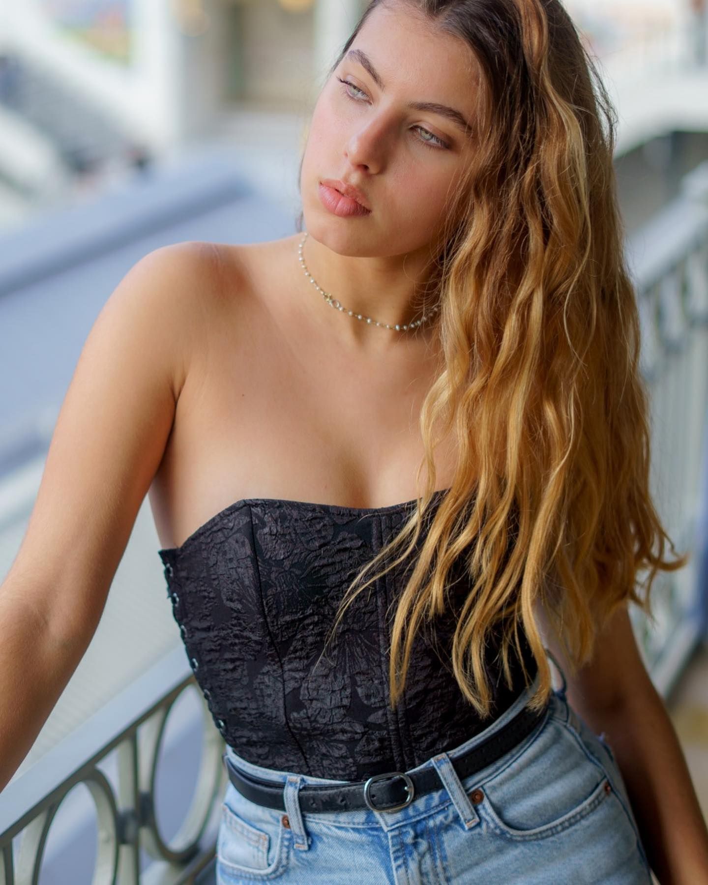 Young woman with wavy blonde hair wearing a black strapless corset top and jeans, posing on a bridge.