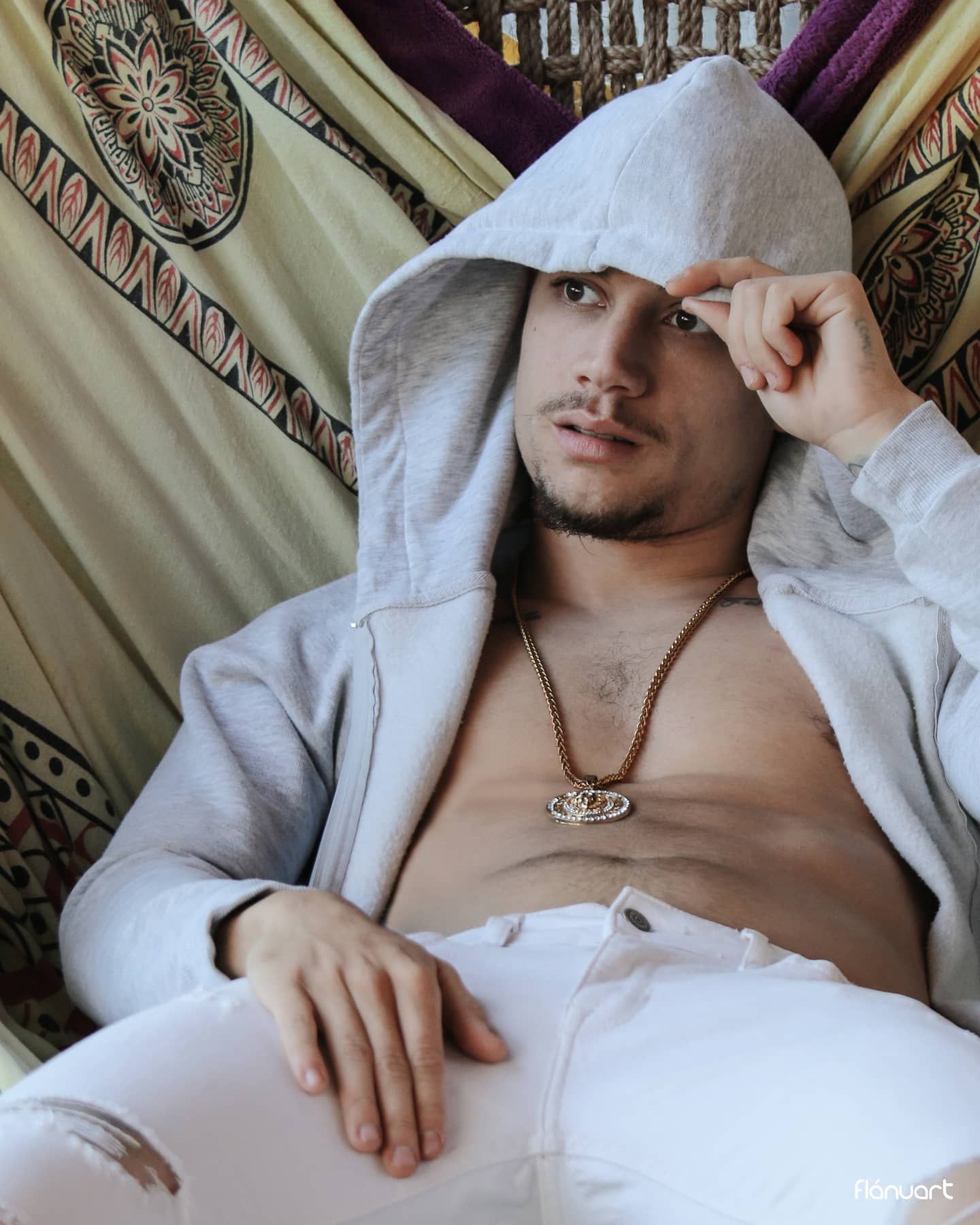 Young man in open grey hoodie and white jeans wearing gold chain necklace, relaxing in hammock.