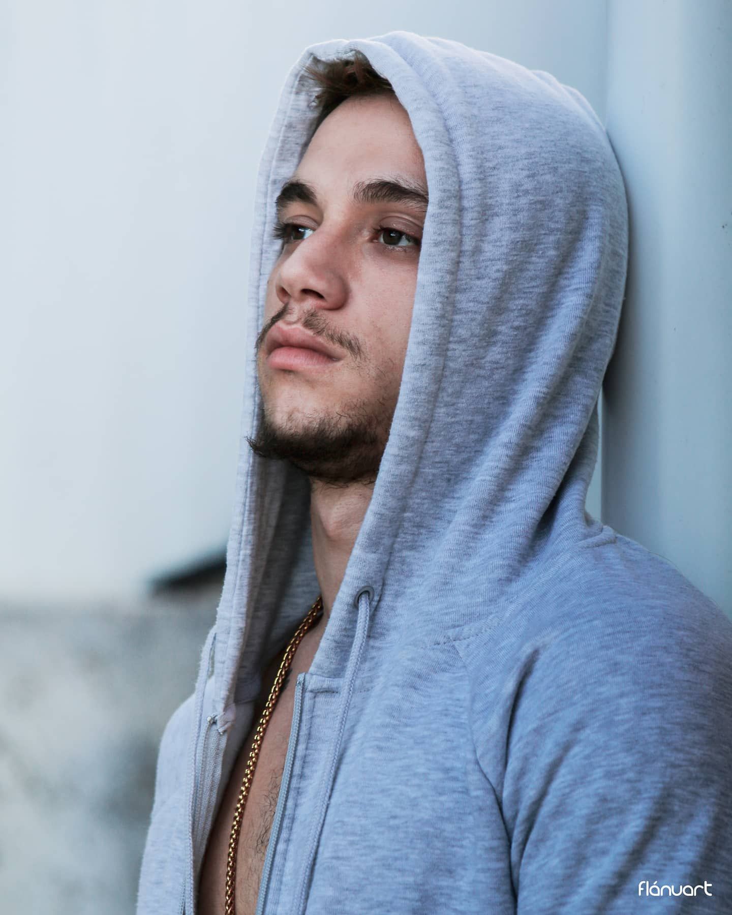 Young man wearing a gray hoodie with a gold chain poses in a moody urban setting.