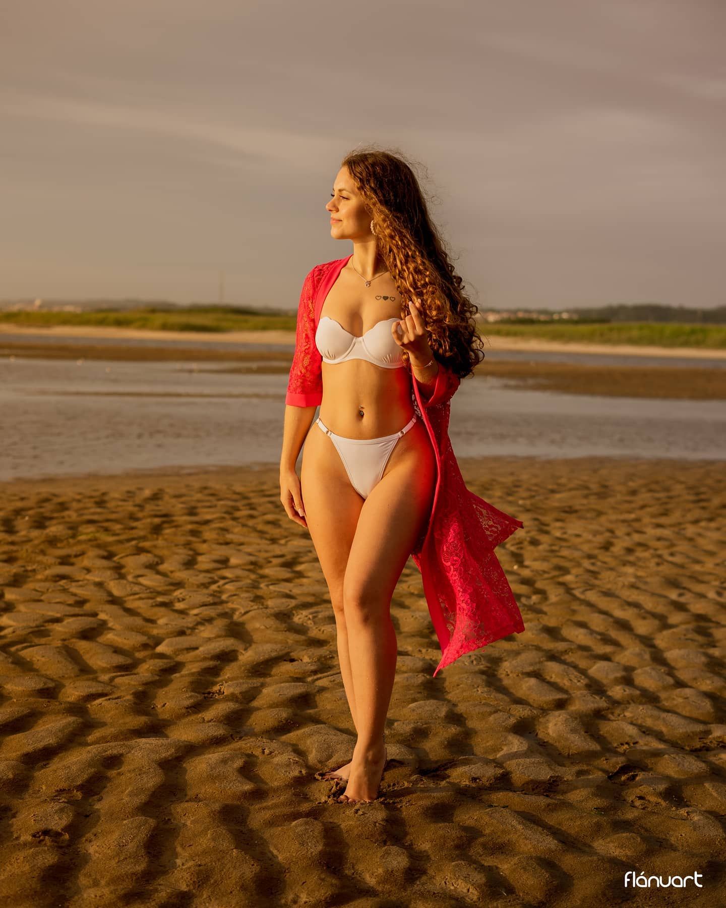 Woman in white bikini with red lace kimono walking on a sandy beach during golden hour.