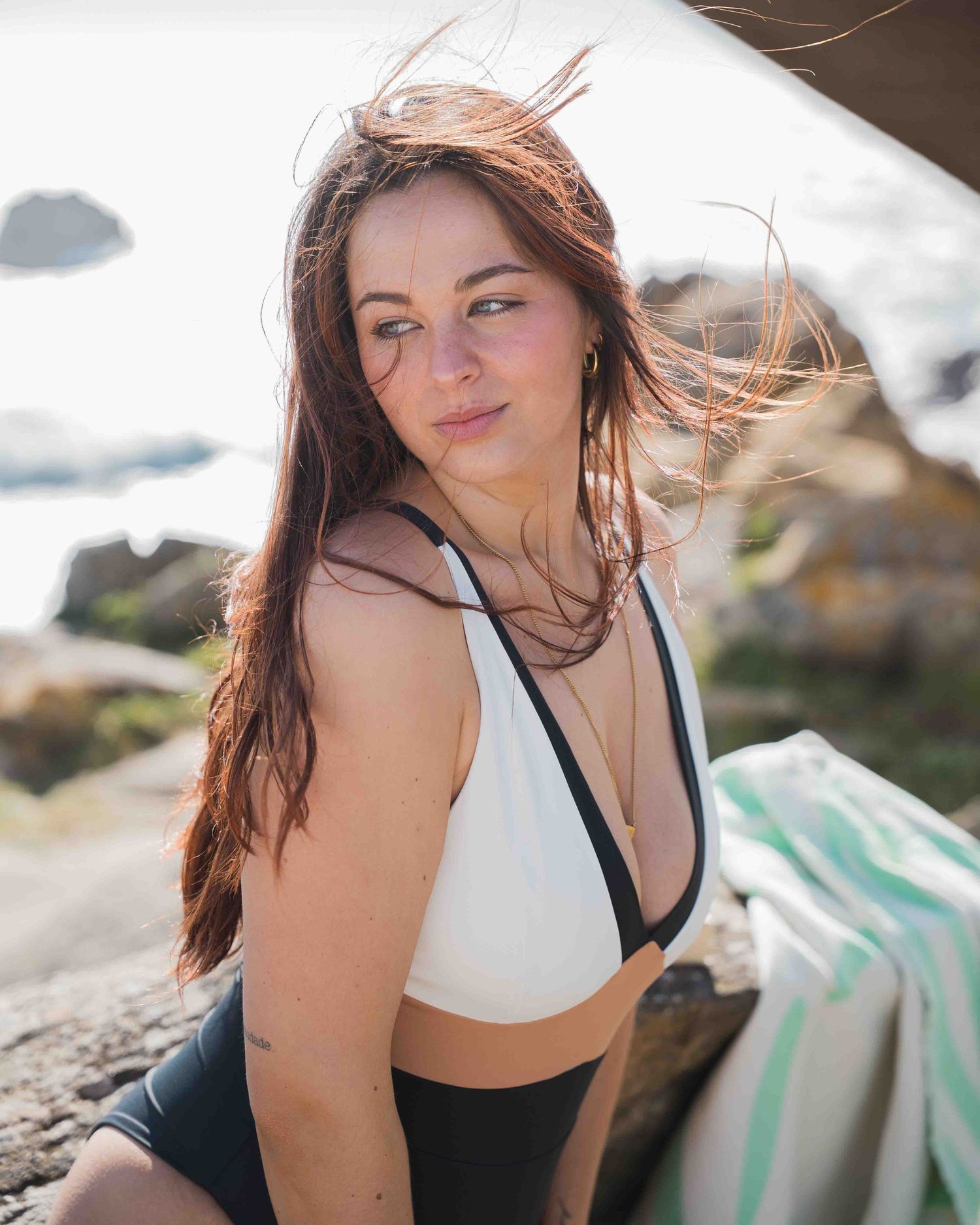 Brunette woman in black and white bikini top posing on rocky beach with ocean backdrop.