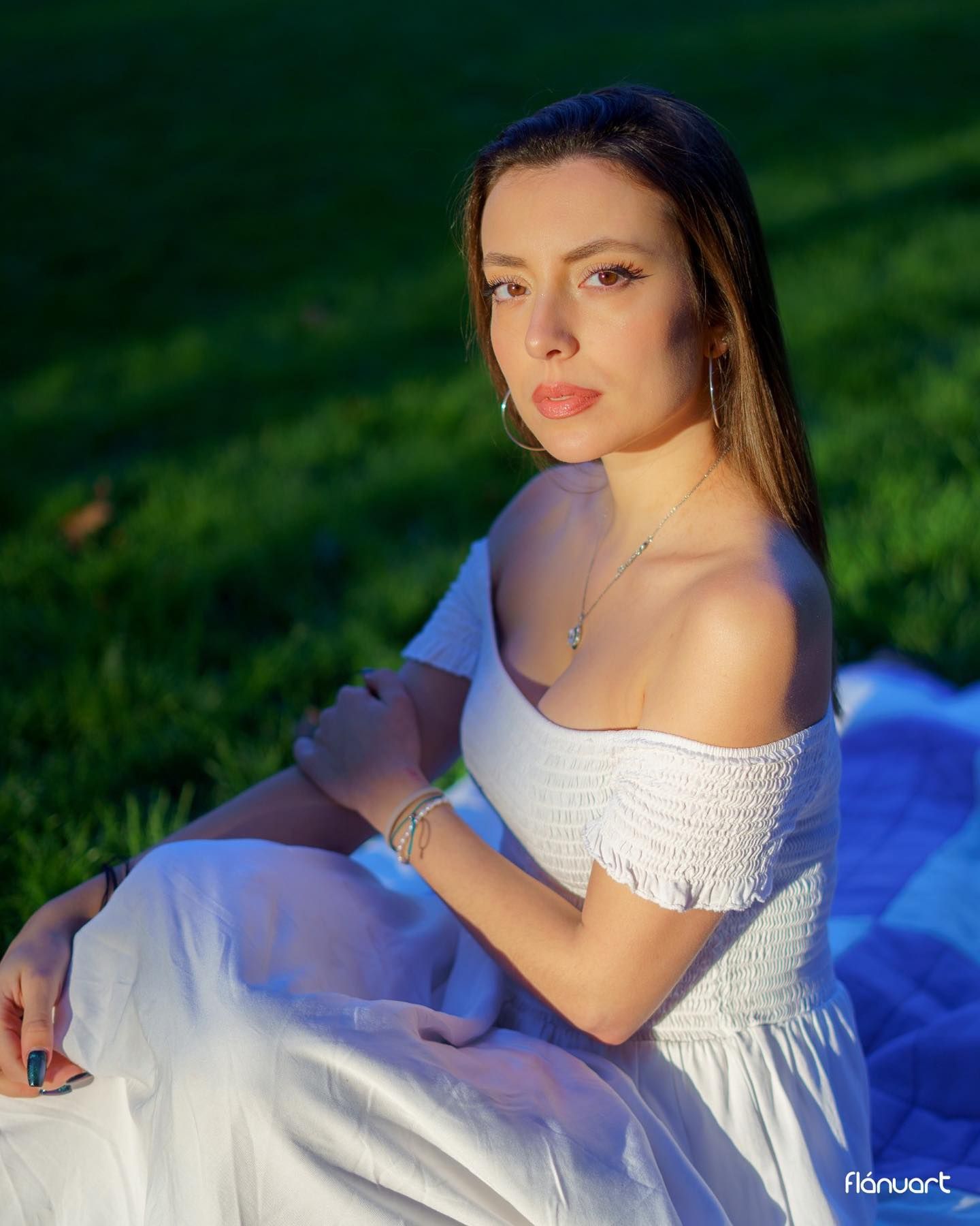 Young woman in white off-shoulder smocked dress sits gracefully on grass in soft evening sunlight.