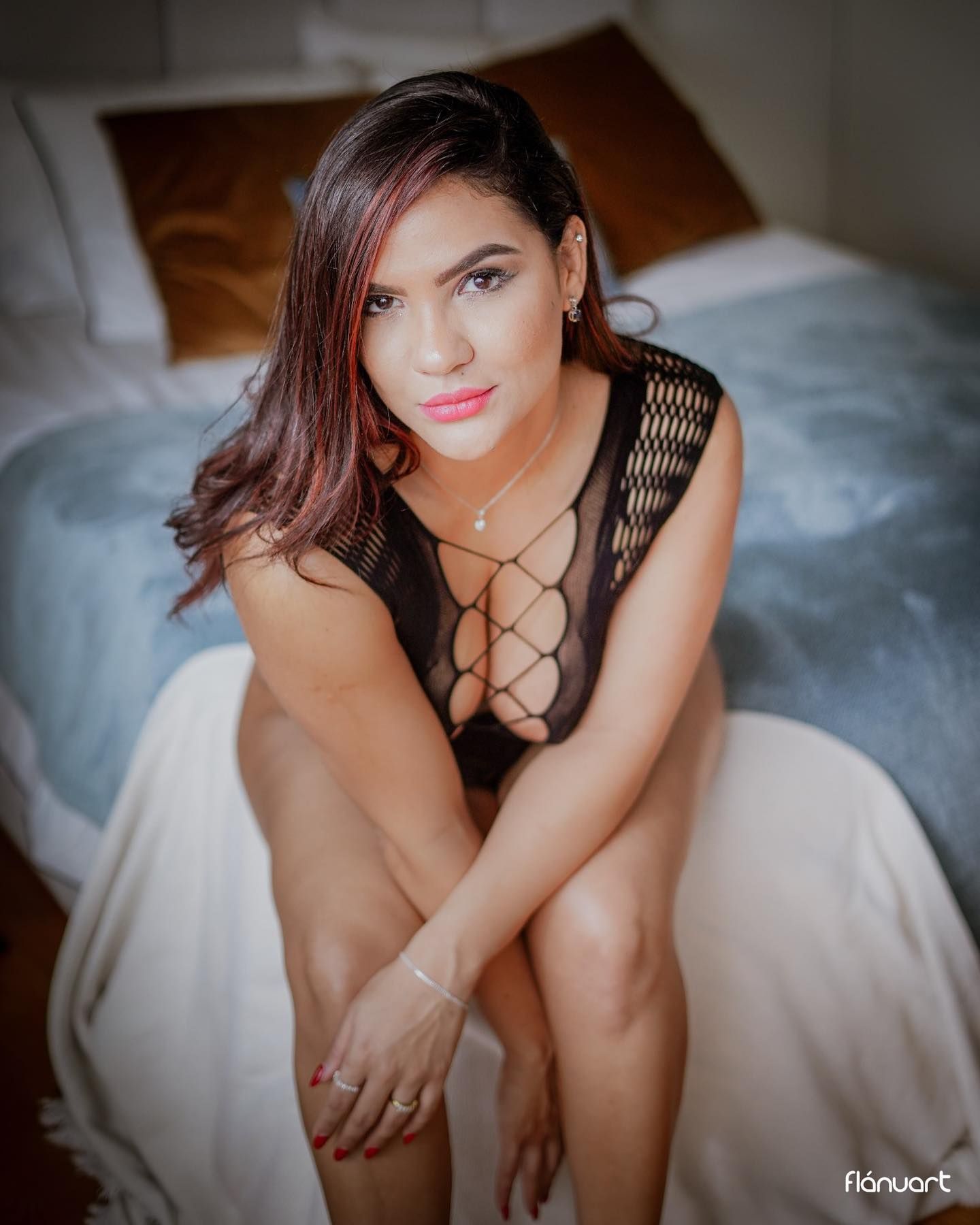 Woman in black mesh bodysuit sitting on white bed, posing confidently for boudoir photo.