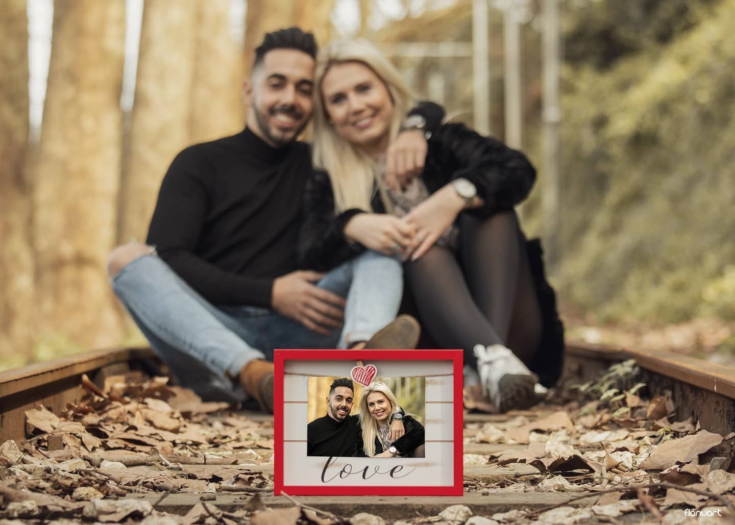 Couple sitting on railway tracks holding a framed love photo surrounded by autumn leaves.
