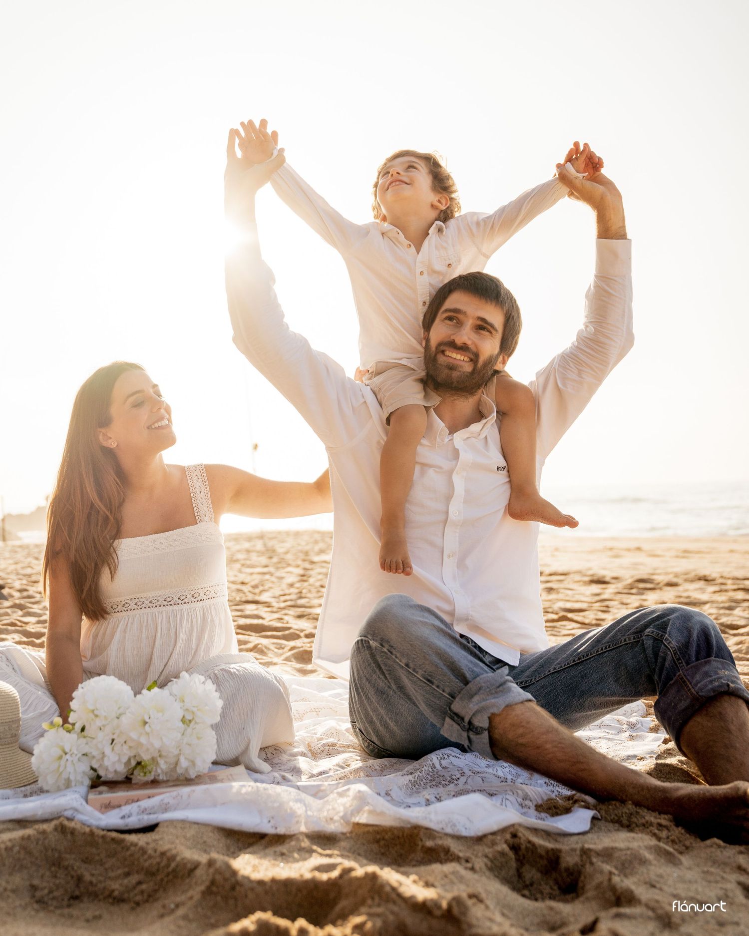 Joyful family at golden hour beach, child sitting on fathers shoulders with arms raised.