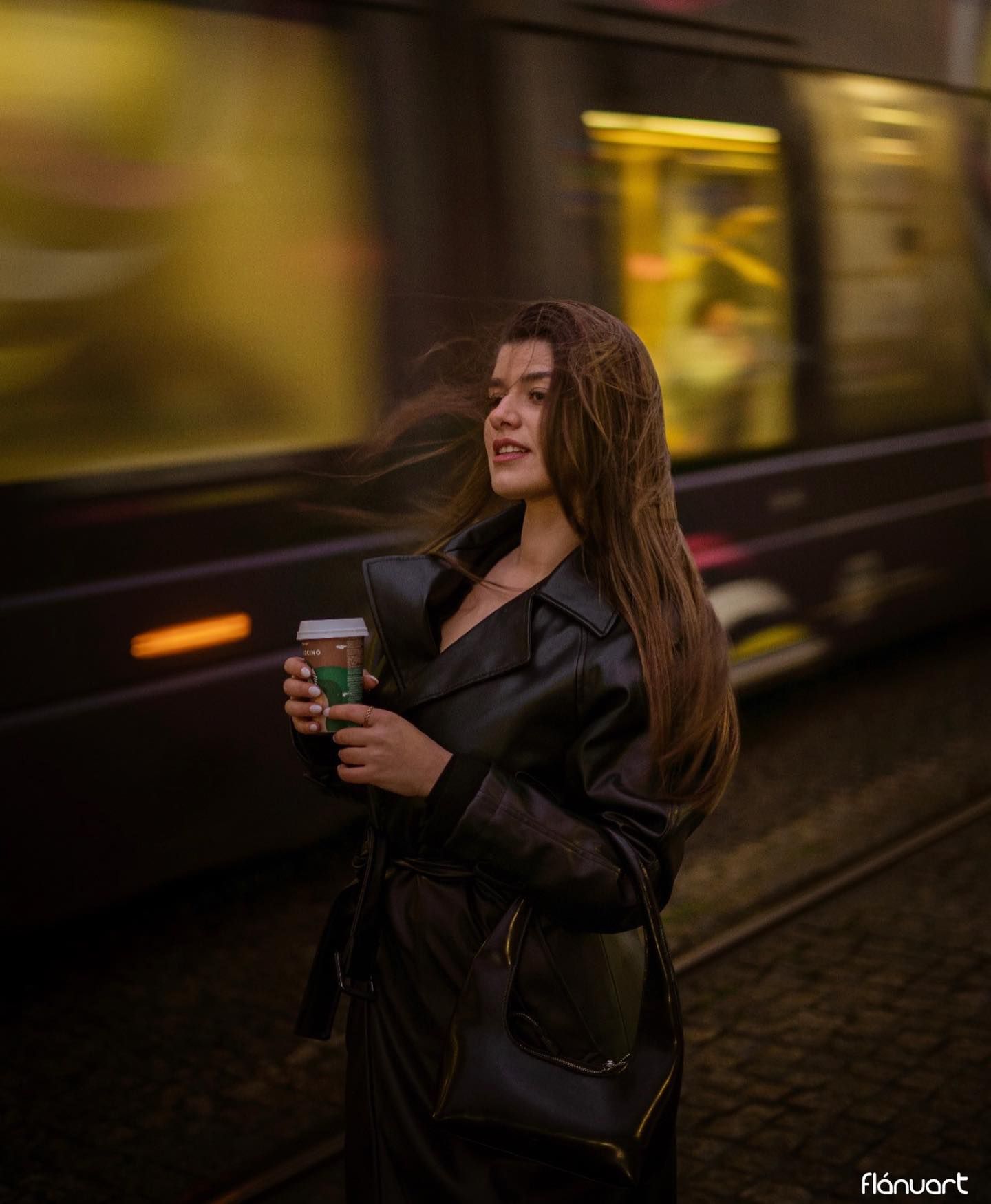 Woman in black leather coat holding coffee cup near passing tram at night.