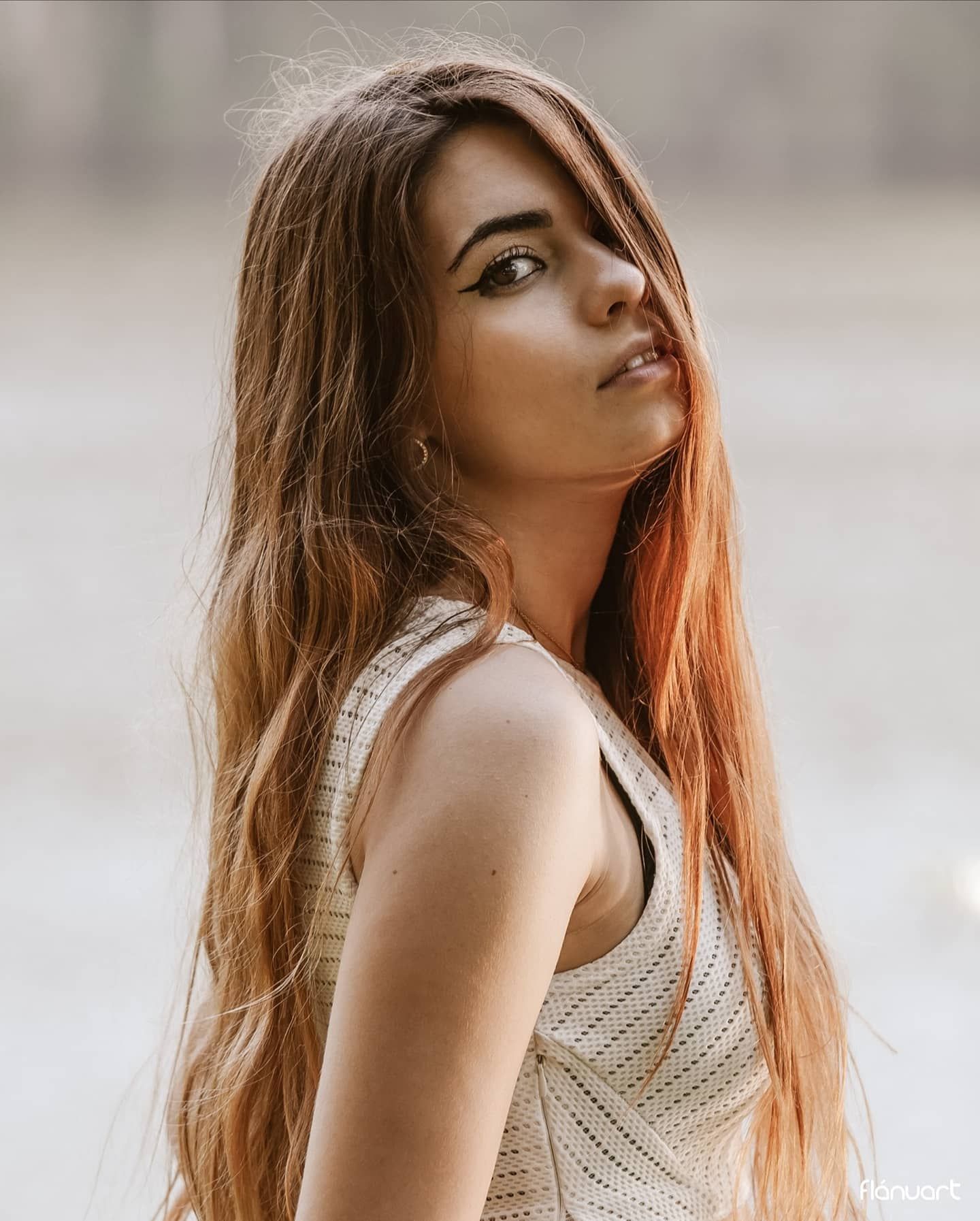 Young woman with long auburn highlighted hair in white knit top posing outdoors with soft bokeh background.