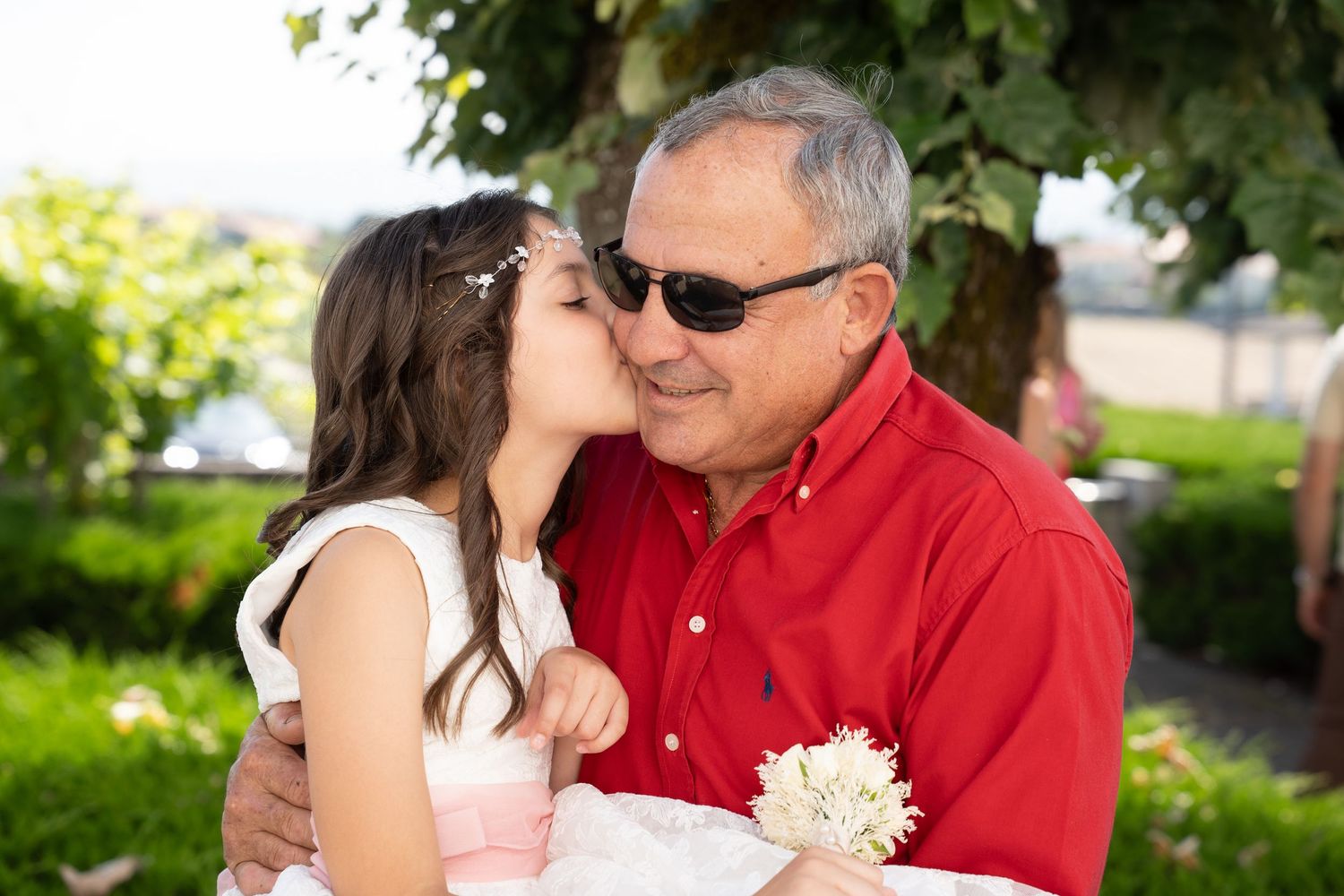 Young girl kissing an older man on the cheek outdoors at a family gathering, man wearing red shirt and sunglasses.