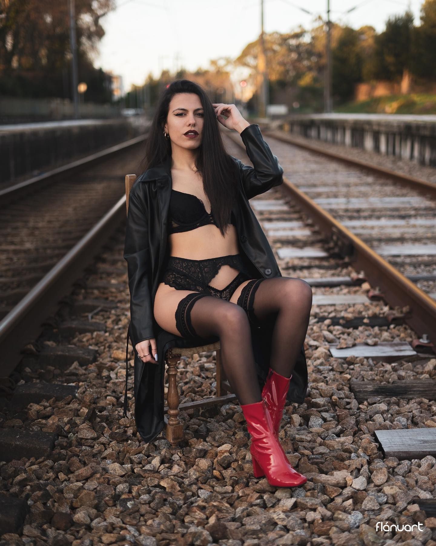 Brunette woman in black lingerie and red boots sitting on train tracks in a leather jacket.