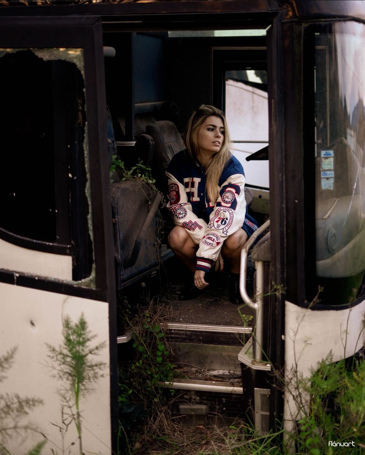 Blonde woman in a varsity jacket sitting inside an abandoned overgrown bus in a moody outdoor setting.