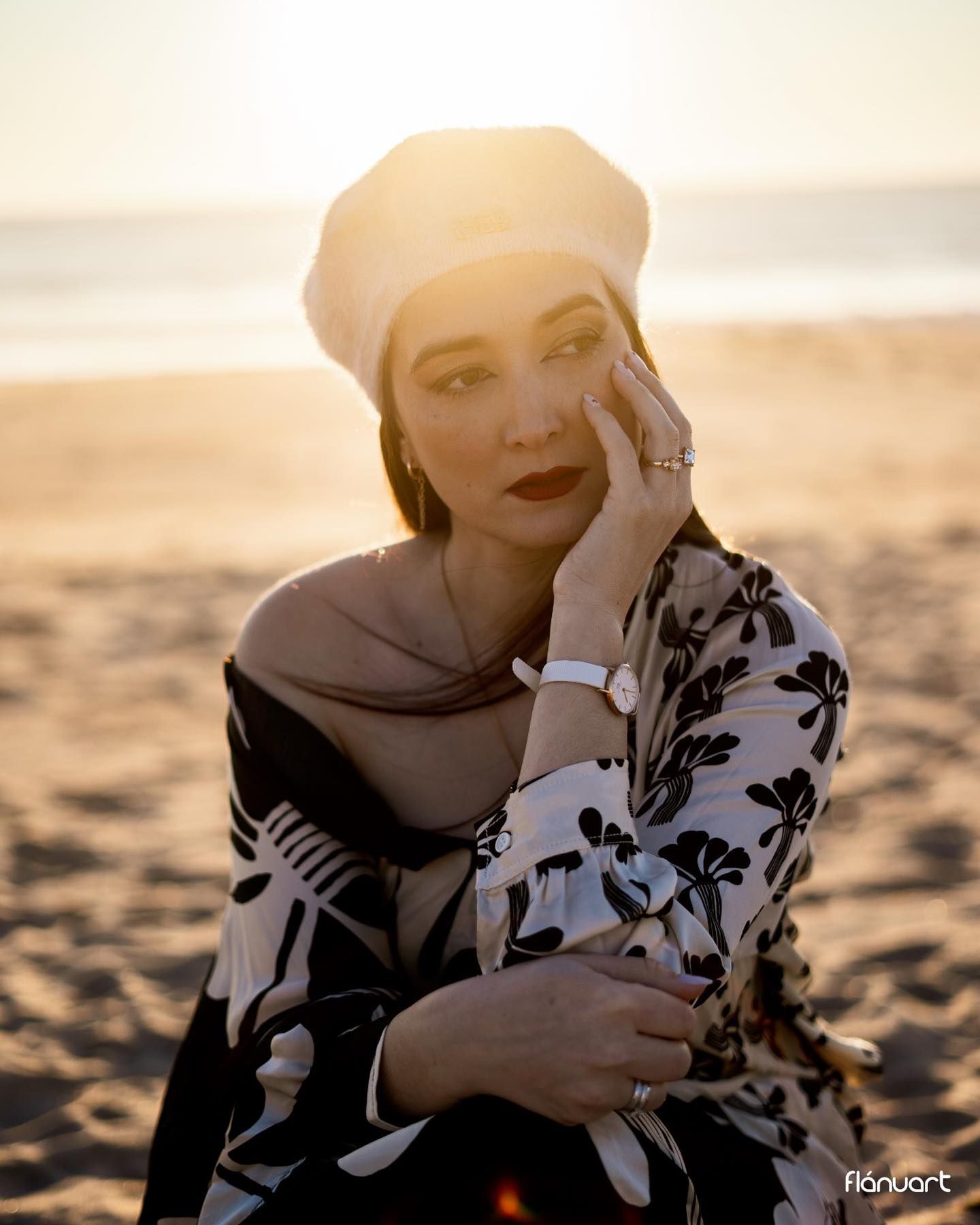 Stylish woman in beret and floral dress posing on beach during golden sunset light.