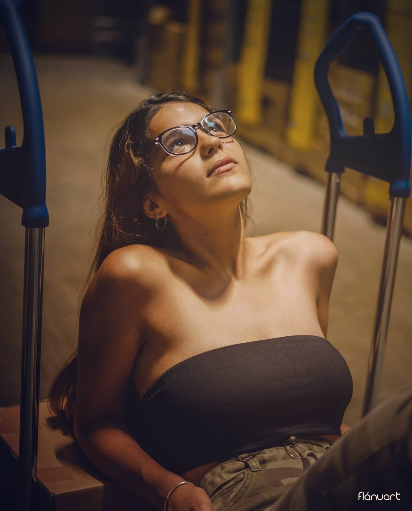 Woman in black tube top tilts head back wearing glasses while seated in warm light.