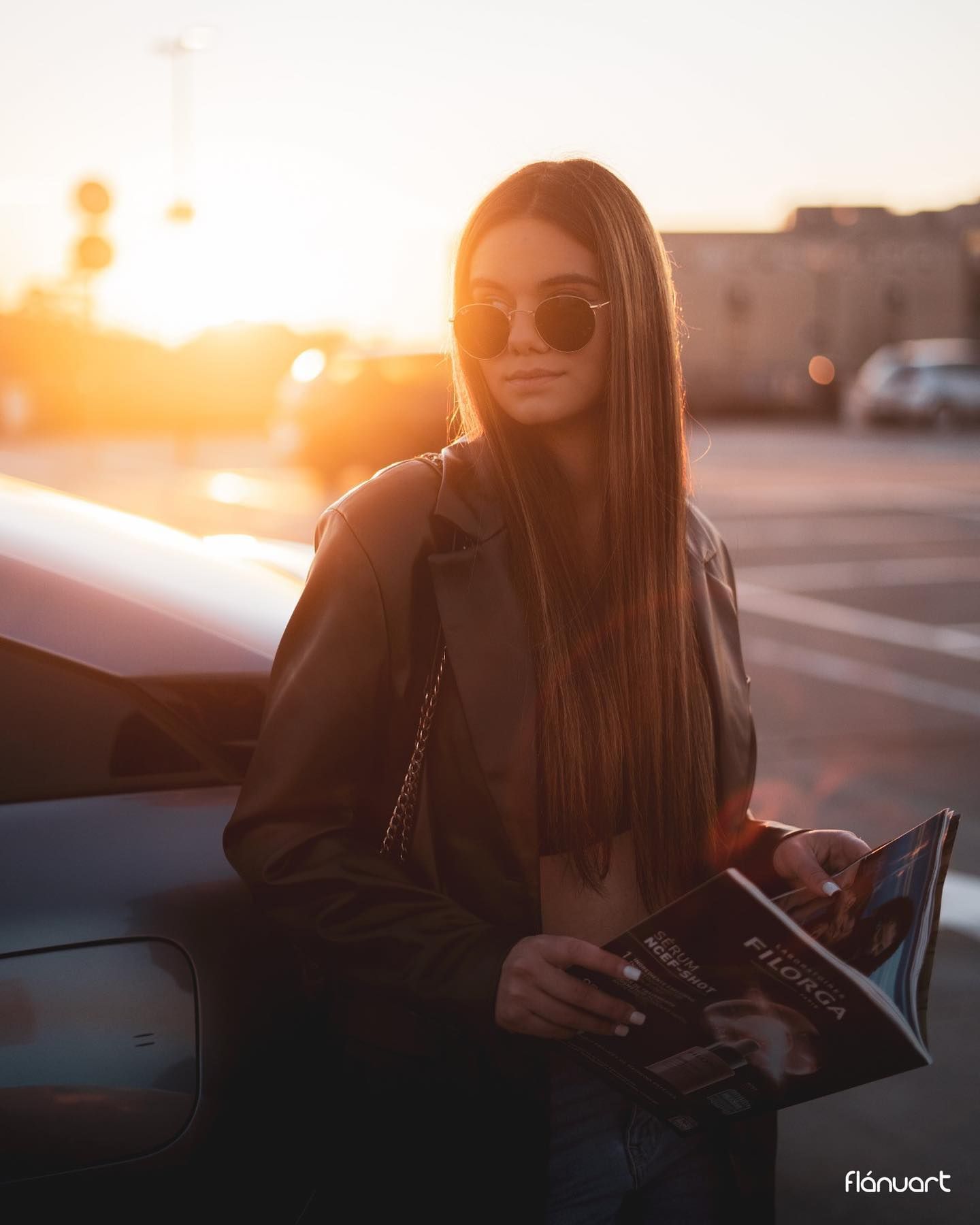 Young woman in leather jacket holding magazine leaning on car during golden hour sunset.