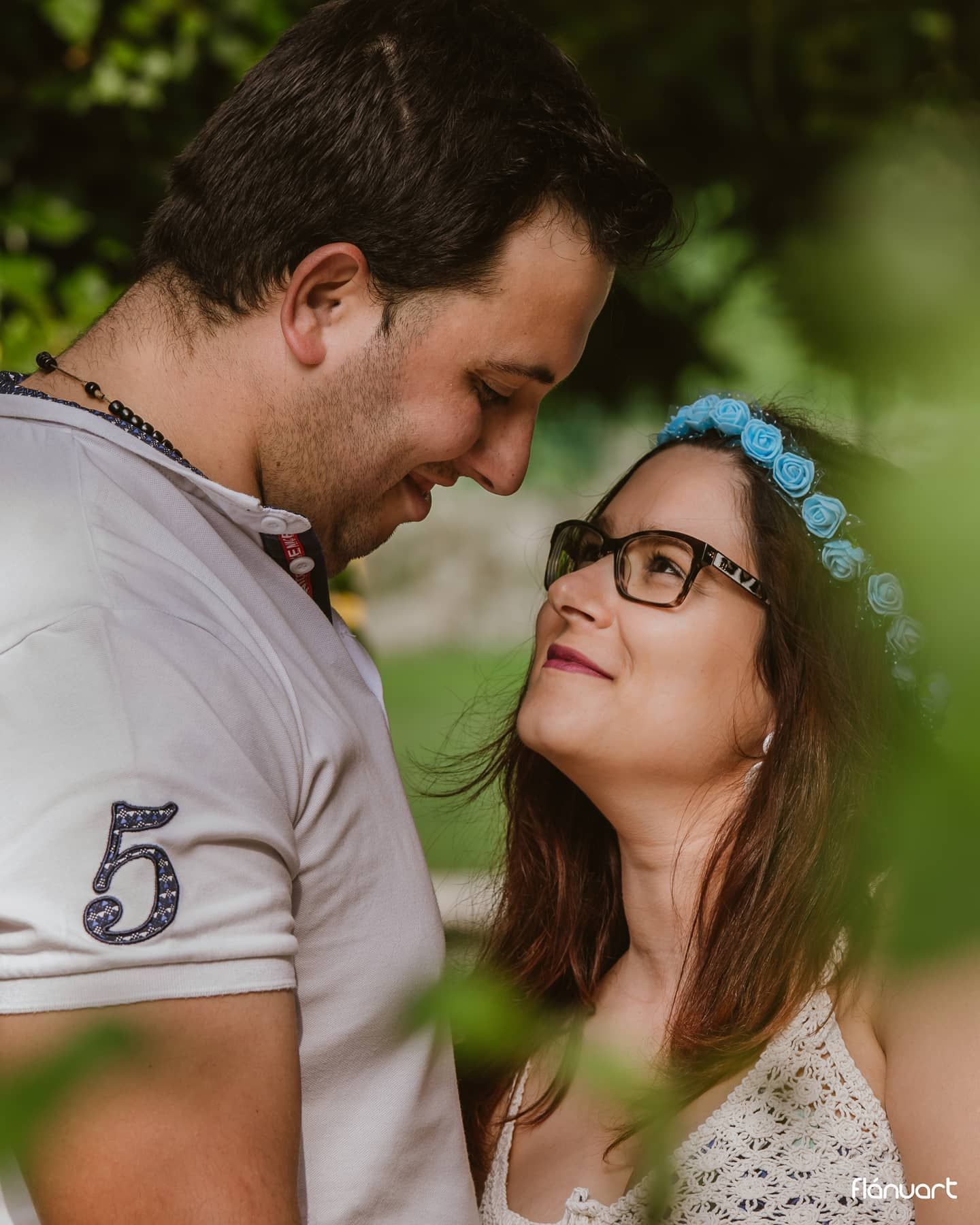 Romantic couple gazing at each other outdoors, woman wearing blue flower crown and glasses, man in white shirt with number 5.
