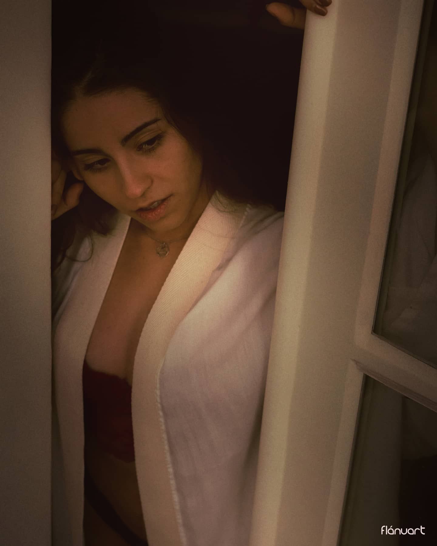 Woman in white robe peering through a window in warm moody lighting.