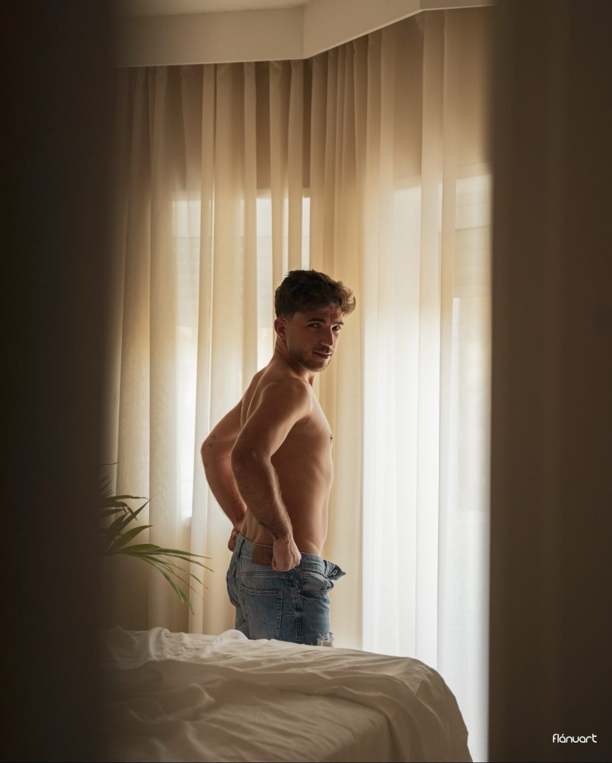 Shirtless athletic man in jeans stands beside a bed in a softly lit room with white curtains.