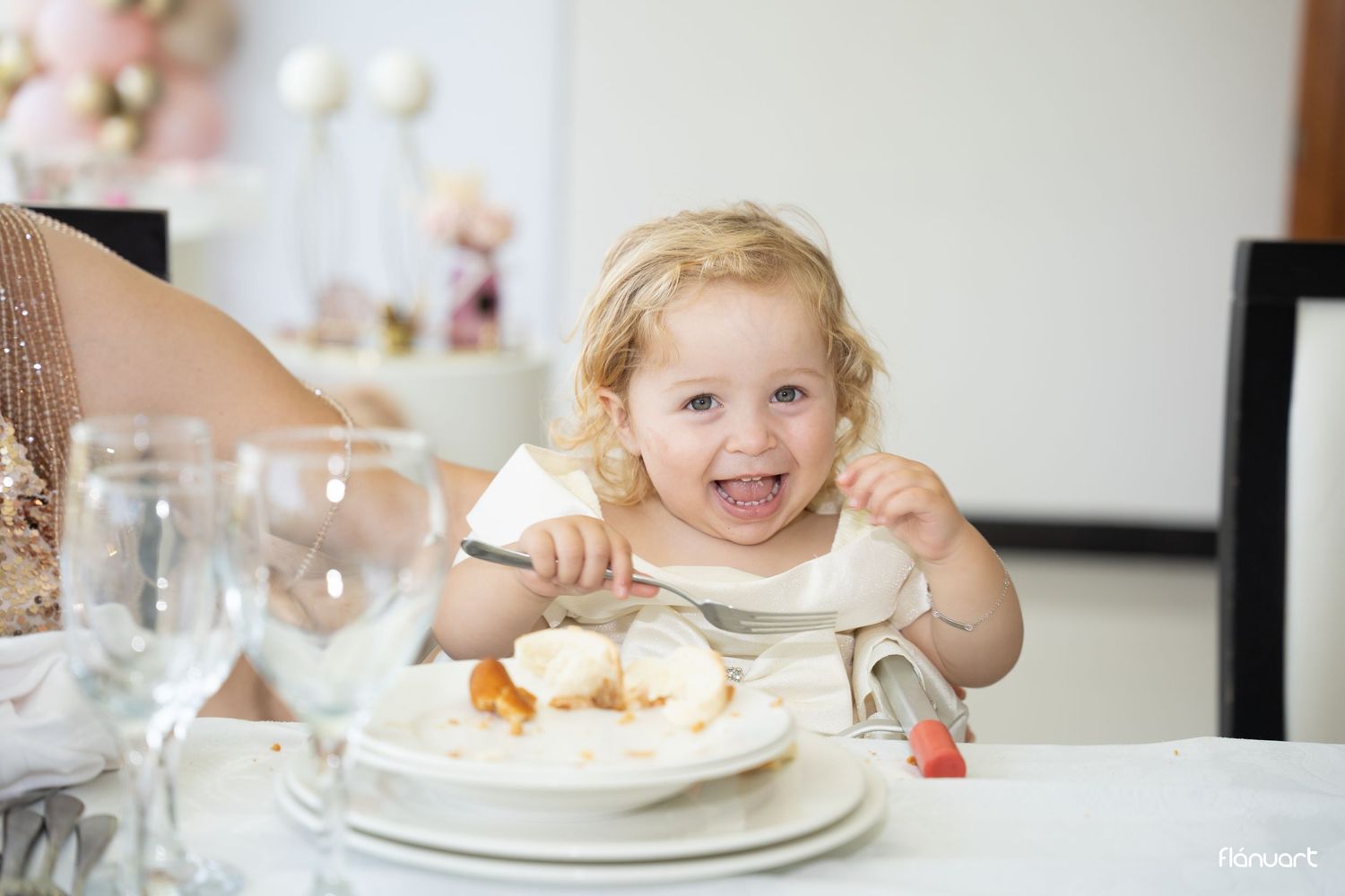 Smiling toddler girl with curly blonde hair eating dessert at a decorated party table.