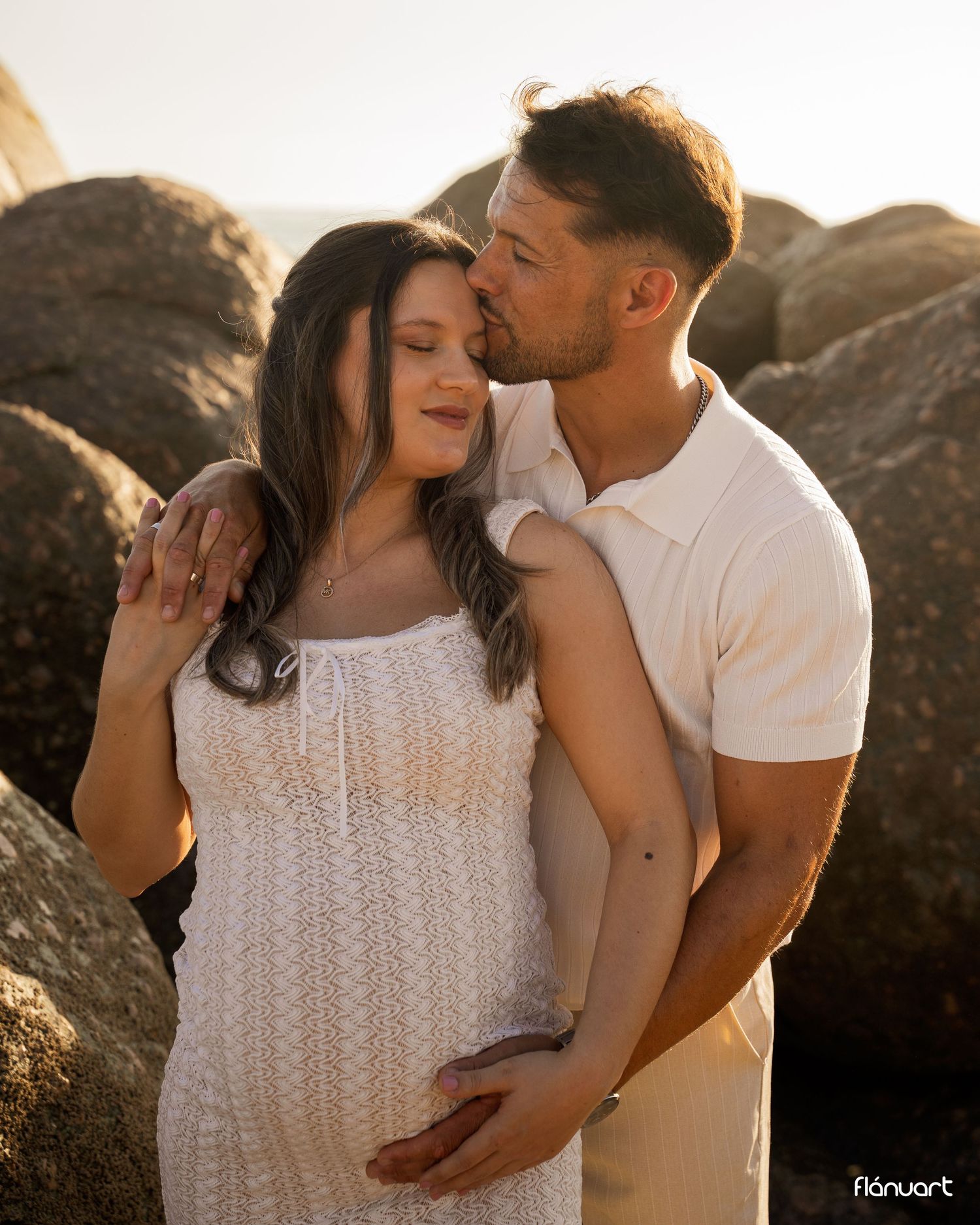 Couple sharing a tender maternity moment on rocky beach at sunset, woman in white lace dress with partner kissing her head.