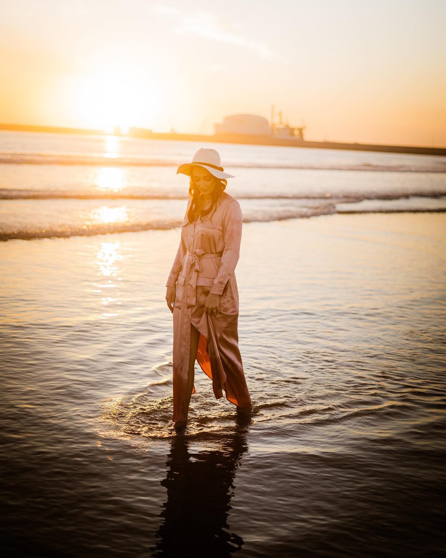 Woman in hat and maxi dress wading in ocean water at golden sunset.