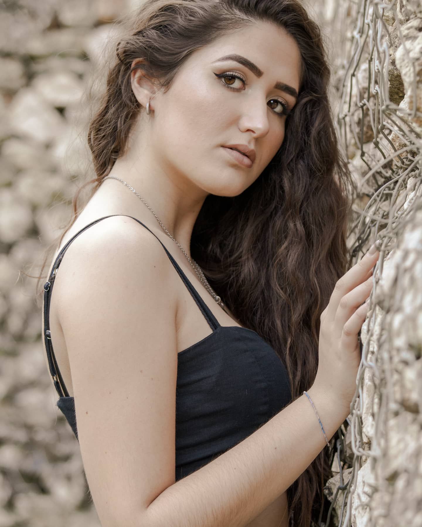 Young woman with long wavy dark hair posing in a black dress against a stone wall outdoors.