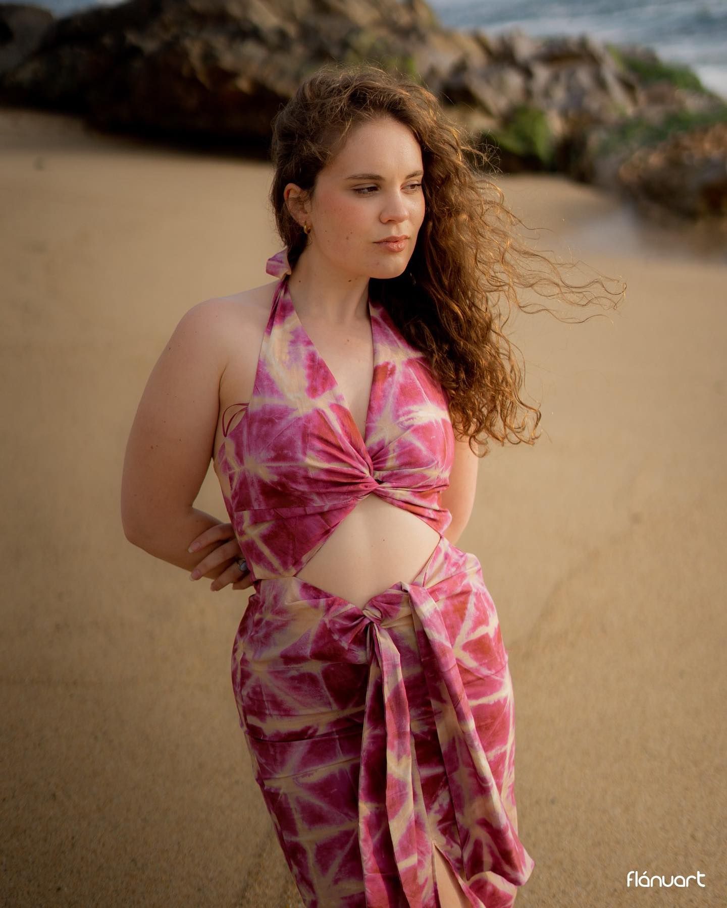 Young woman in pink tie-dye beach outfit standing on sandy shore with rocky backdrop.