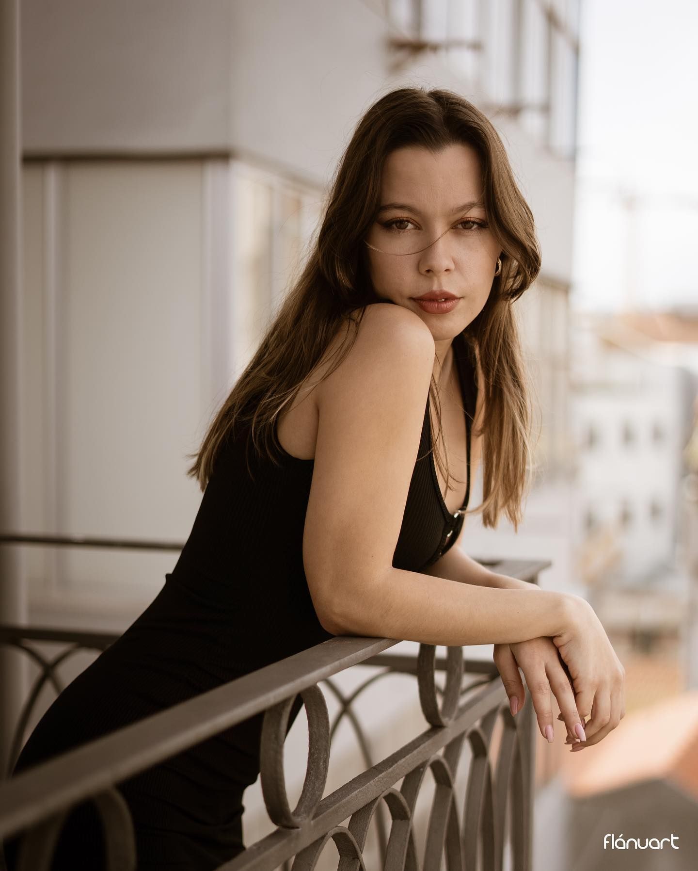 Young woman in black dress leans on balcony railing with urban backdrop in soft light.