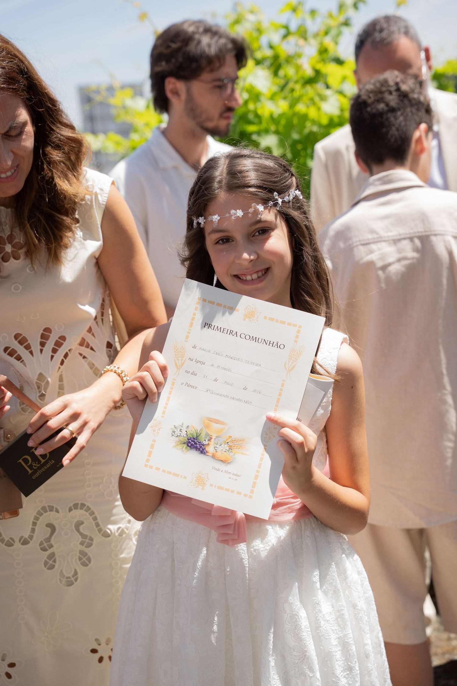 Young girl in white dress proudly holds First Communion certificate outdoors, smiling with family gathered behind her.