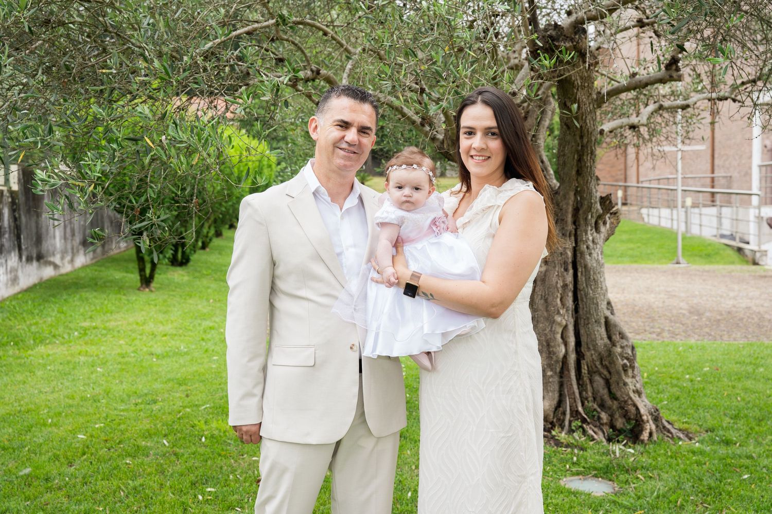 Family portrait of couple holding baby girl in white dress, standing under olive tree on green lawn.