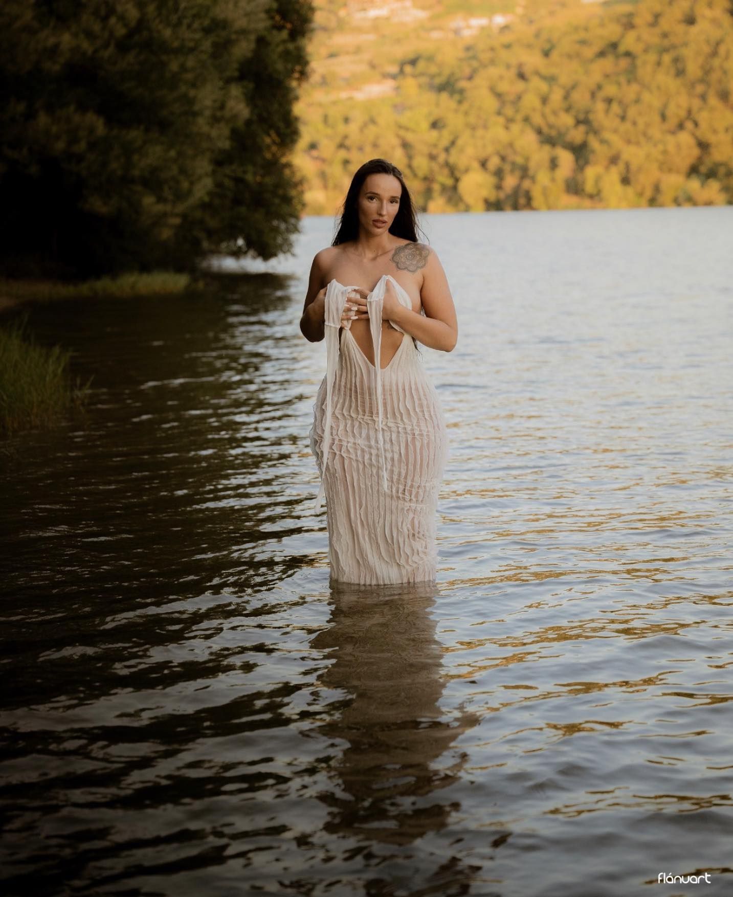 Woman in sheer white dress standing in lake water surrounded by lush green forest scenery.