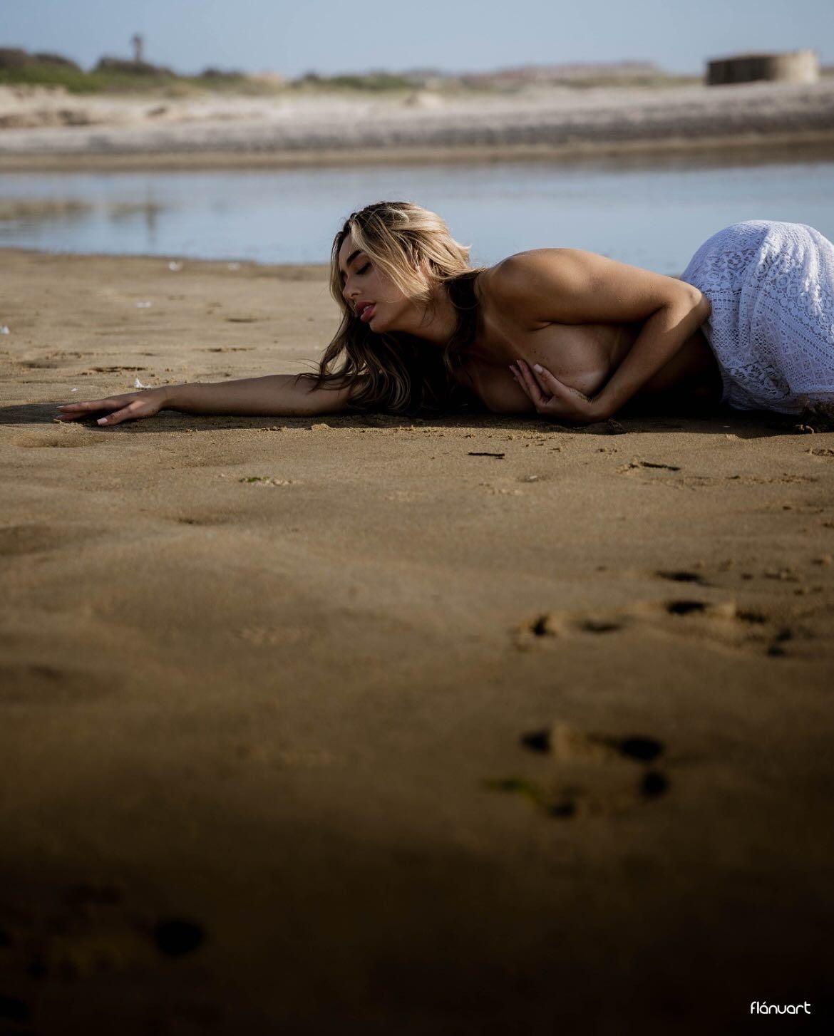 Blonde woman in white dress lying gracefully on sandy beach with coastal water background.