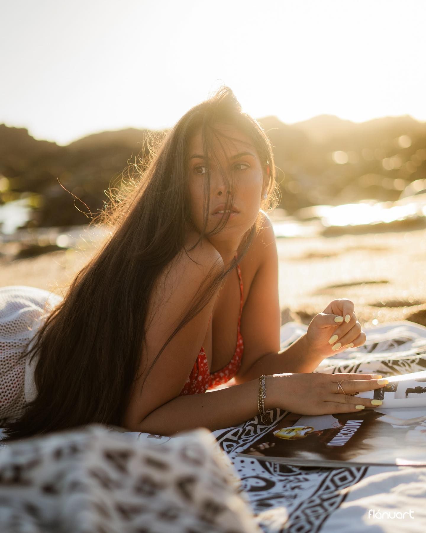 Woman with long dark hair lying on patterned blanket on beach in red bikini at sunset.