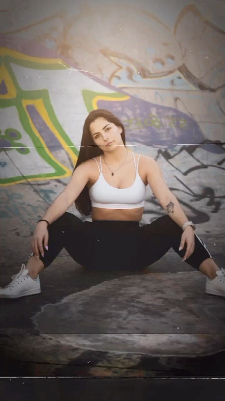 Athletic woman in white sports bra and black leggings sitting in splits before graffiti wall.