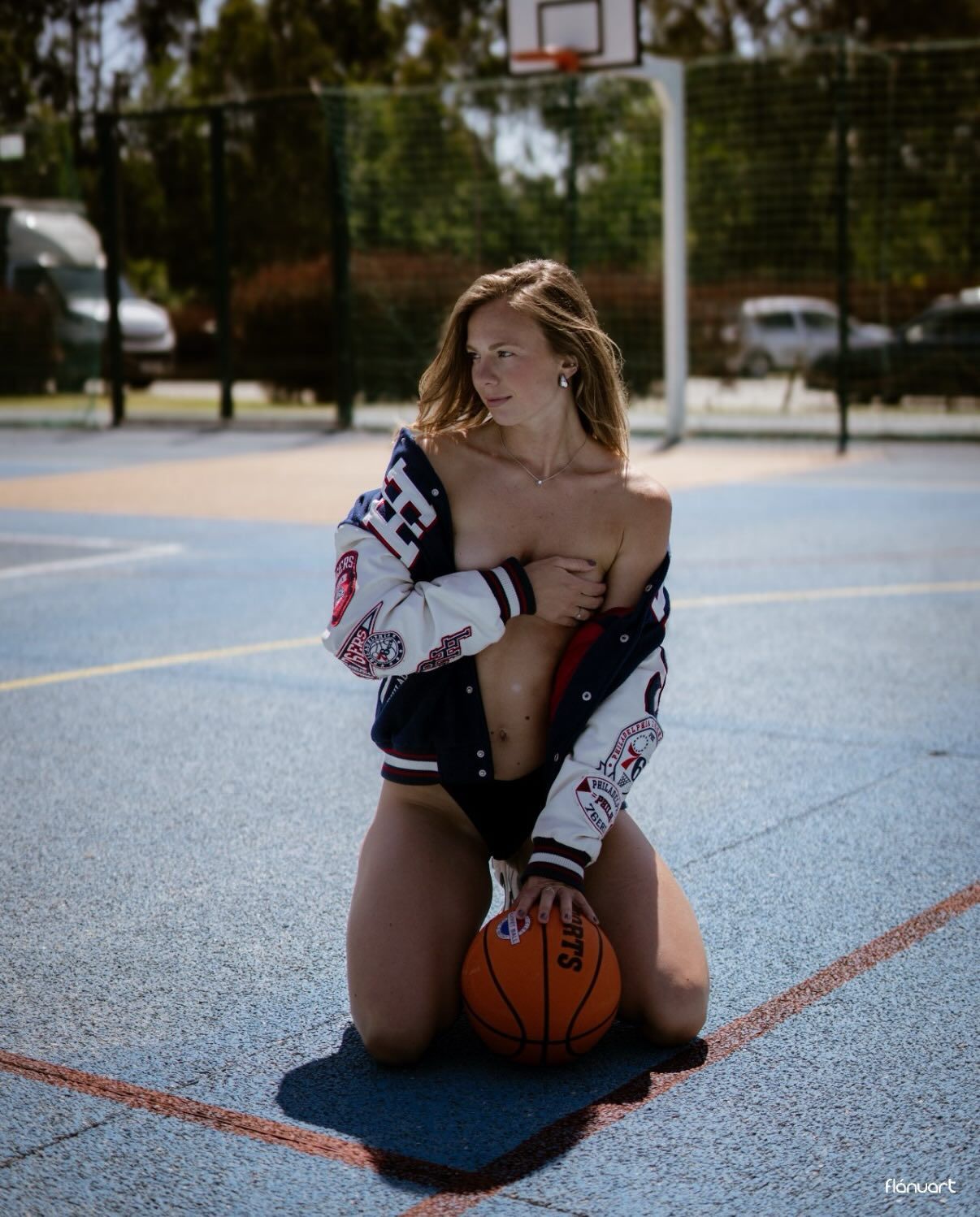 Blonde woman in open varsity jacket kneels on outdoor basketball court holding a basketball.