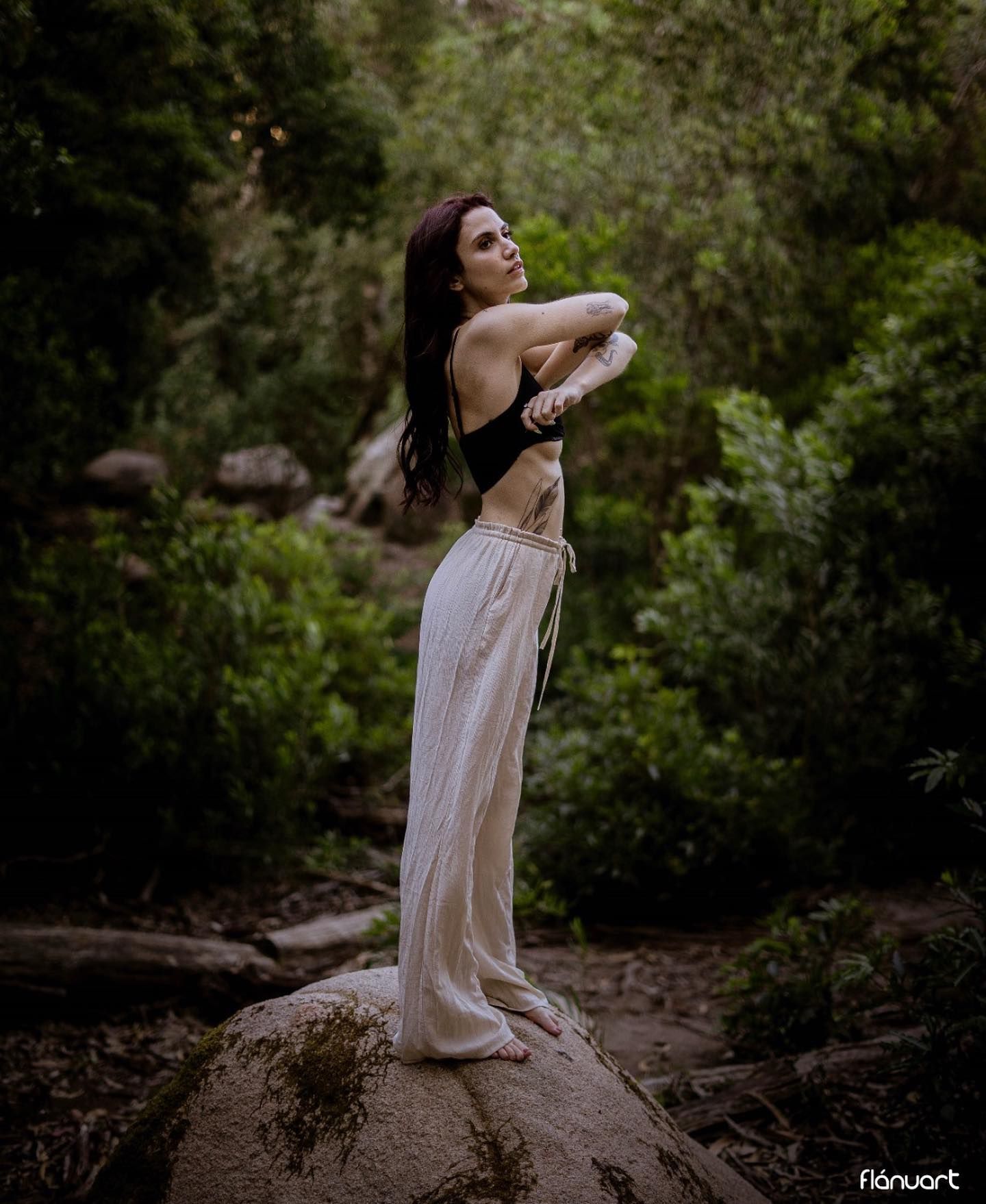 Dark-haired woman in black crop top and white pants posing on rock in forest.