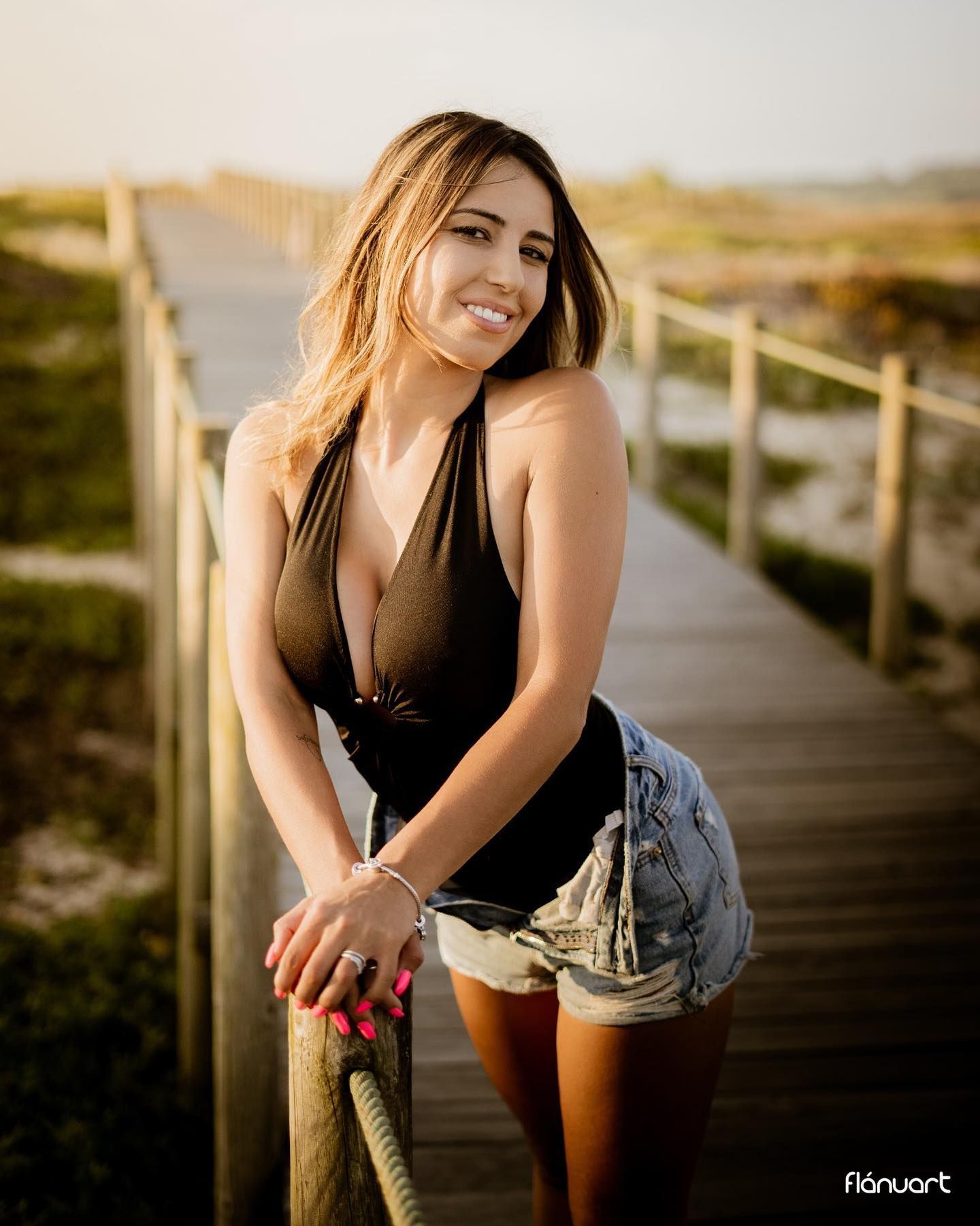 Smiling blonde woman in black swimsuit and denim shorts posing on a wooden boardwalk at sunset.