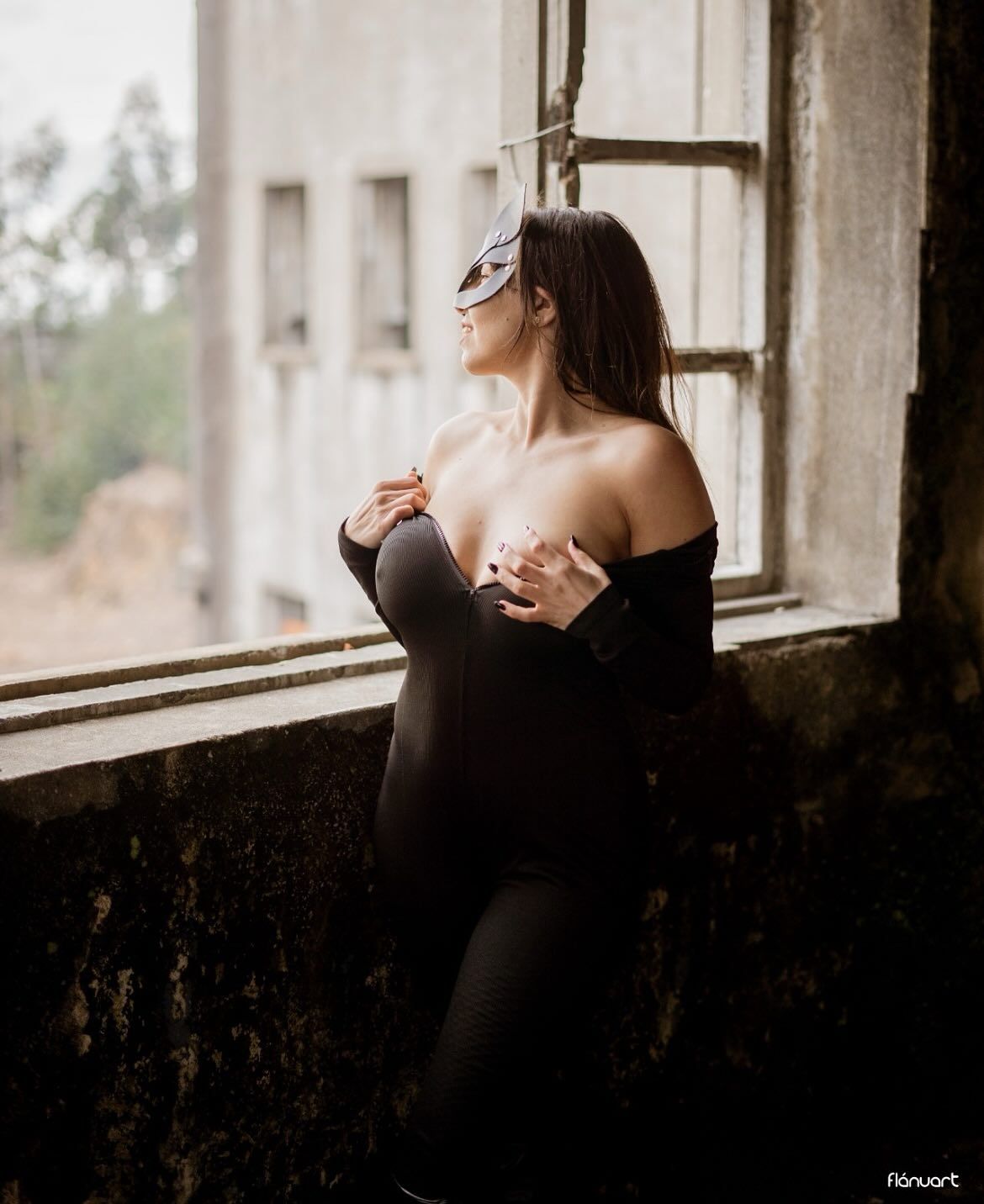 Woman in black bodysuit and masquerade mask posing by a rustic window in an abandoned building.