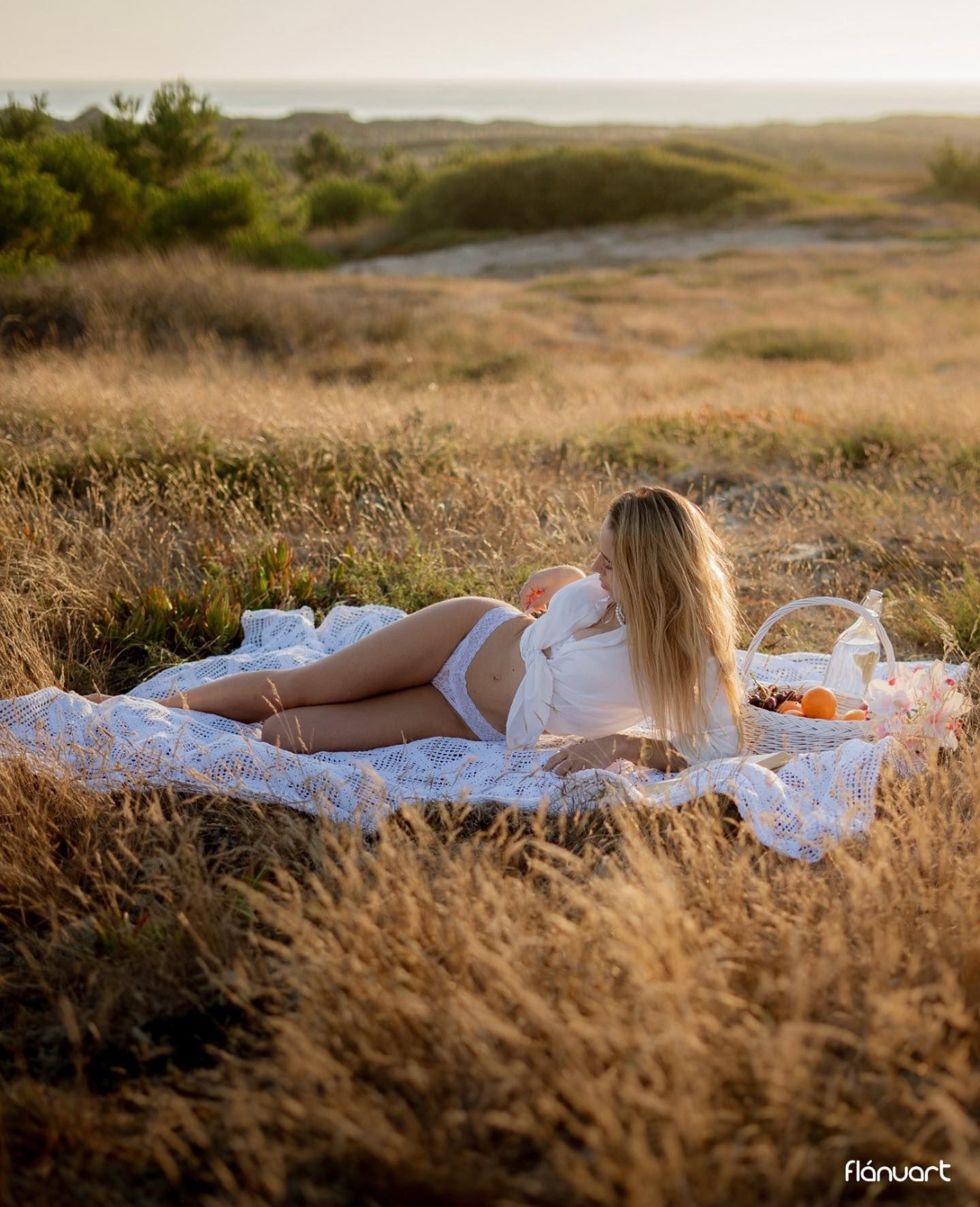 Blonde woman in white lingerie relaxing on a lace blanket in a golden field at sunset.