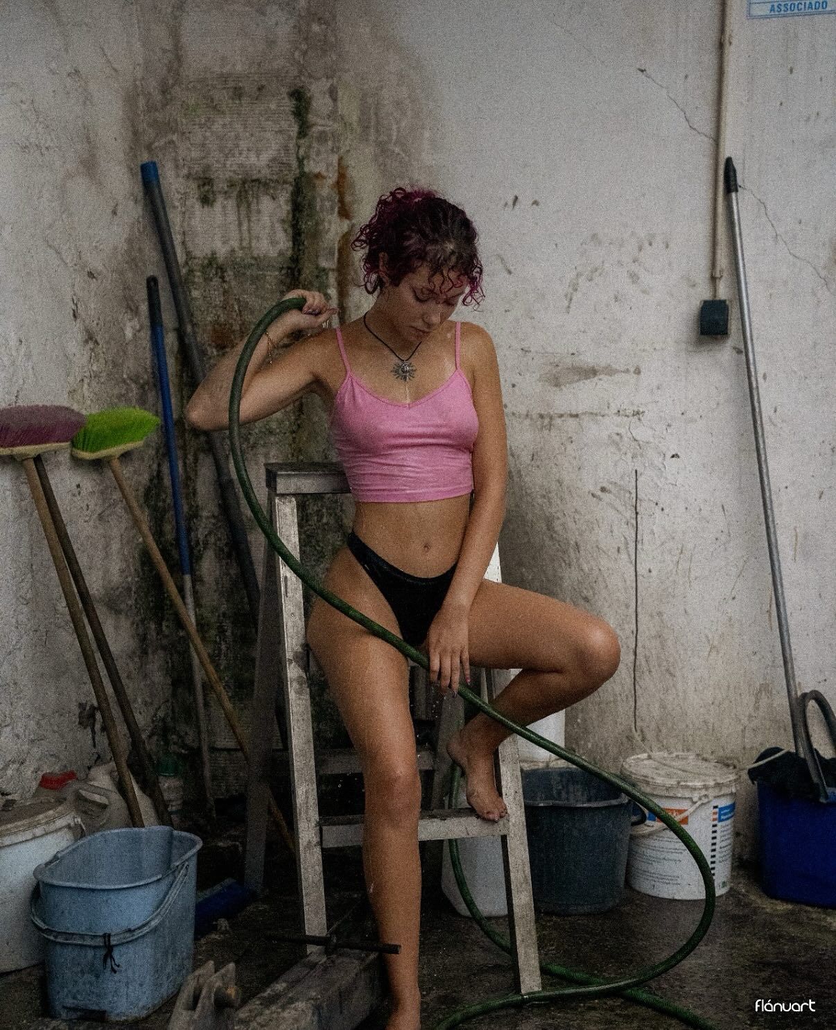 Woman with red curly hair in pink crop top and black shorts holds a garden hose in a rustic space.