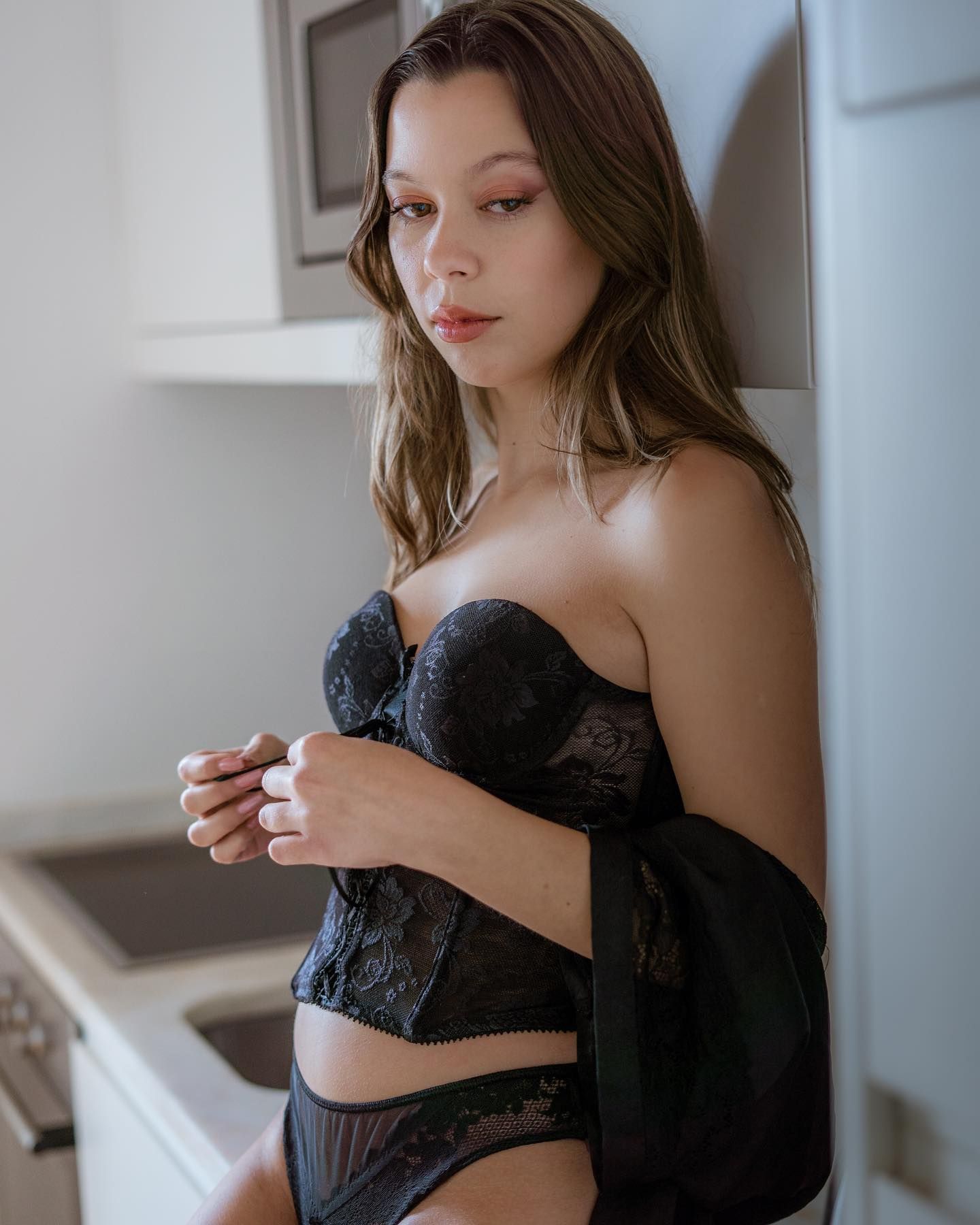 Woman in black lace strapless corset lingerie leaning against a white wall.