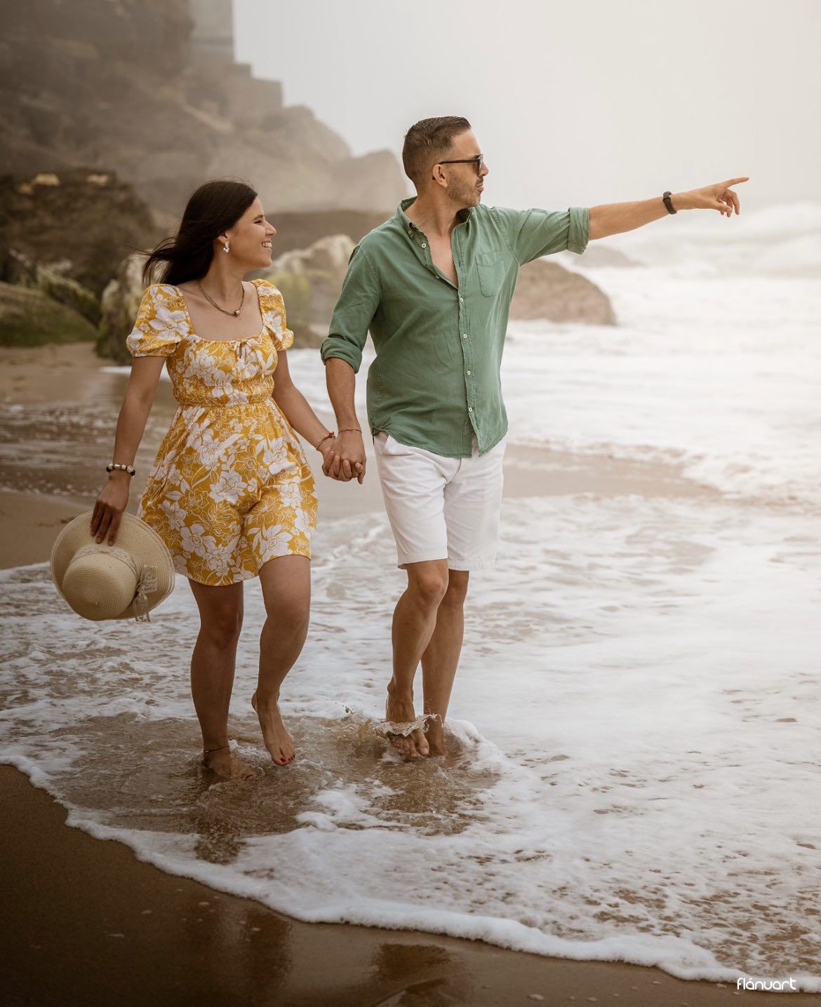 Couple walking along shoreline, man in green shirt pointing into distance while holding woman's hand.