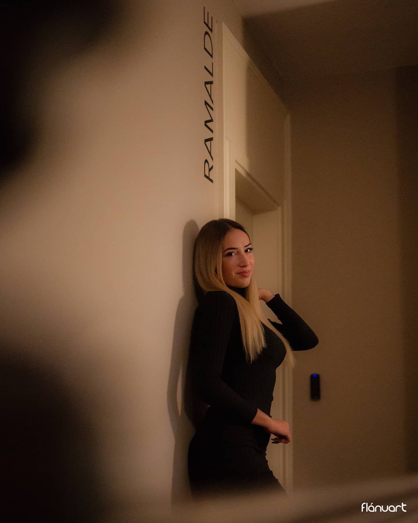 Blonde woman in black outfit leans against wall near Ramalde sign in dimly lit interior.